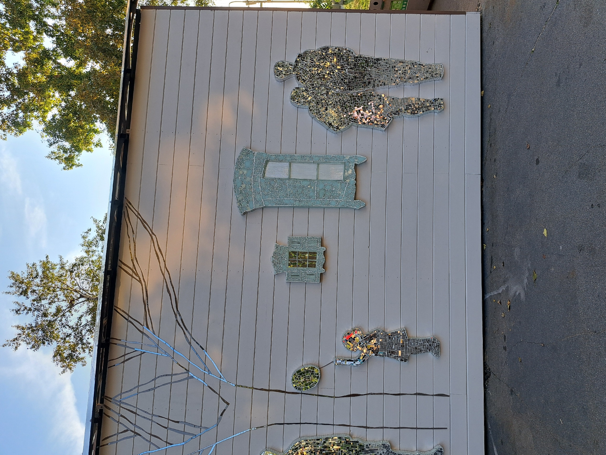 This image depicts a mural on the side of a light-colored, horizontally paneled building. The mural features several mosaic figures made of mirrored tiles. On the left, there is a mosaic silhouette of a person. To the right of this, a child mosaic figure holds a mosaic balloon. Further to the right are two mosaic silhouettes of people standing close together. Above the figures, there are two mosaic window frames, one smaller and one larger, with intricate designs. A mosaic tree trunk and branches extend from the top left of the mural across the building's facade. The building is situated outdoors, with a dark asphalt surface in the foreground, scattered with a few fallen leaves. Trees with green and autumnal foliage are visible in the background, against a bright blue sky. The overall scene suggests an outdoor public art installation. The building appears to be part of a structure like a shed or small building, possibly in a park or a public space. The style of the mosaic art and the presence of a large tree suggest a temperate climate, possibly autumn given the leaves. Without more information, identifying the specific location in Irkutsk, Russia is not possible from the image alone.