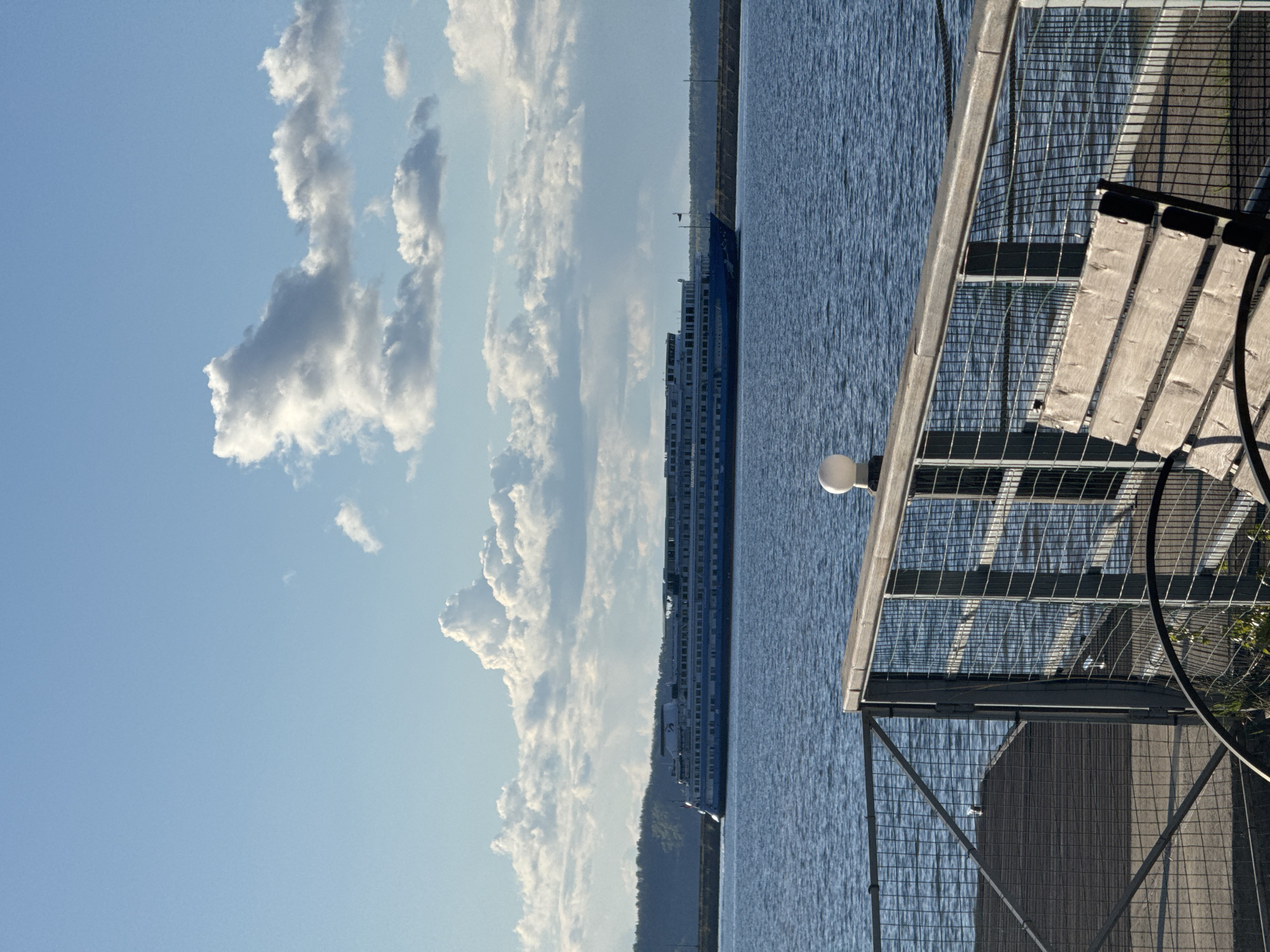 The image shows a view from a pier in Chaikovsky, Russia, with a large ferry boat on the water. The pier has a metal railing and a wooden bench. In the background, there are clouds in the sky, and land can be seen on the horizon. The water appears calm and the sky is blue.