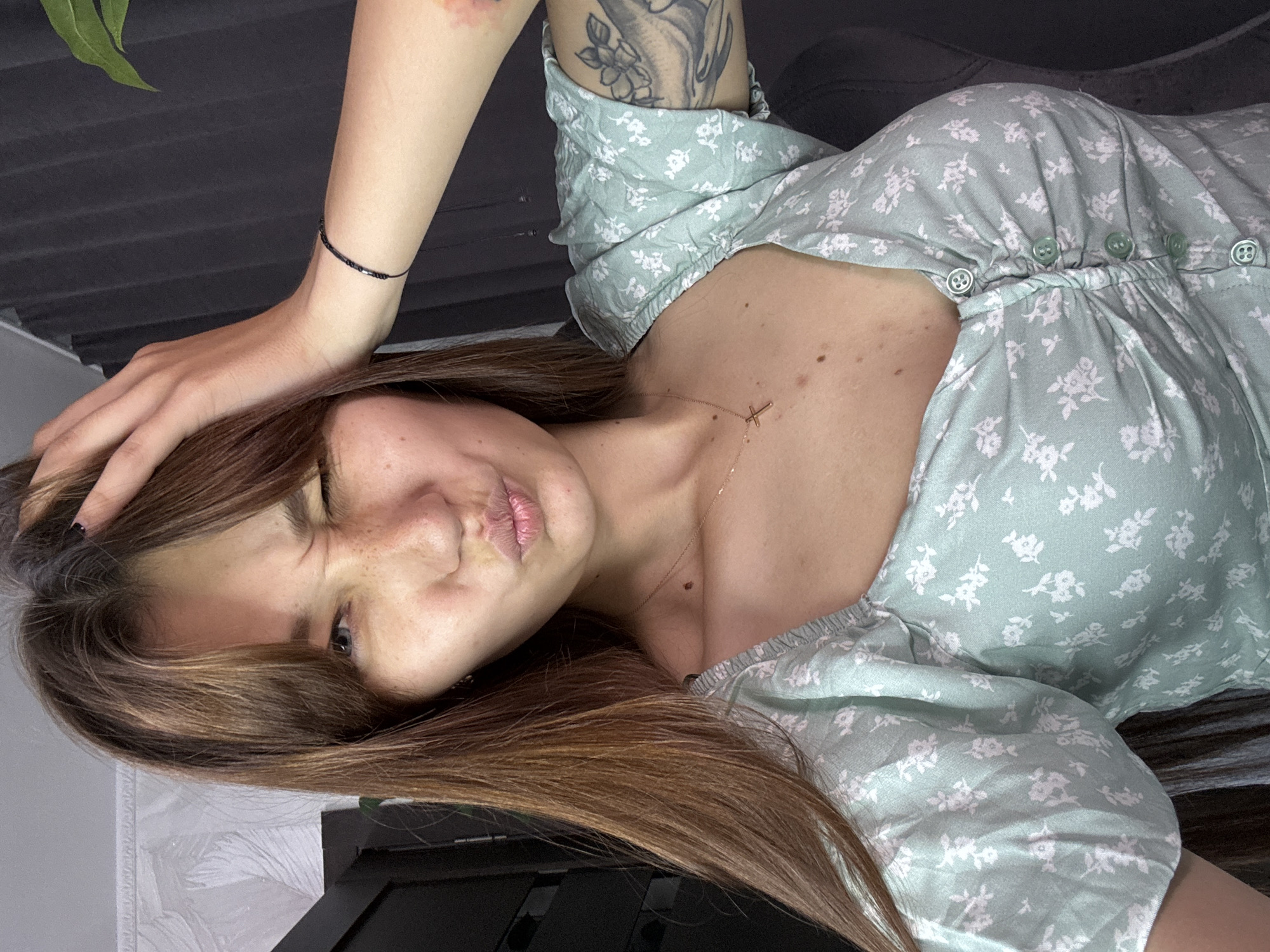 A young woman with long brown hair and freckles is pictured, winking and puckering her lips. She is wearing a light green floral print dress with a ruffled neckline and buttons down the front. A thin black beaded bracelet adorns her left wrist. A cross necklace hangs around her neck. Her right arm is bent, with her hand on her head. A tattoo is visible on her right arm. The background is a dark, out-of-focus wall. The image appears to be taken indoors, possibly in a residential setting. There is no indication of the city of Novokuznetsk, Russia.