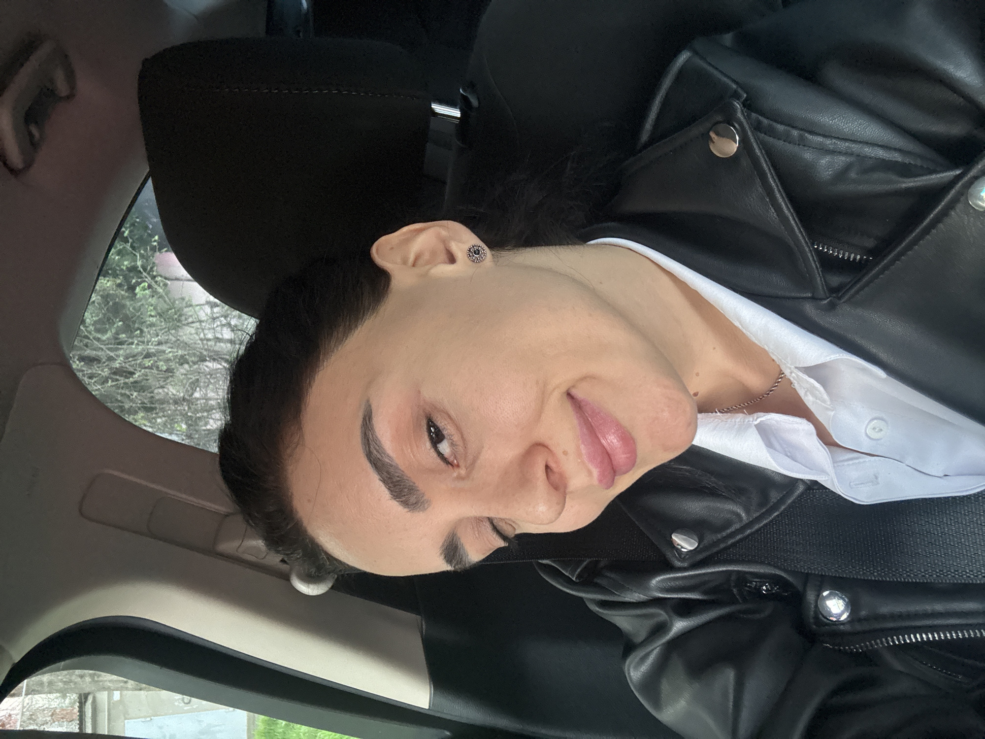 A person with dark hair and light skin is sitting in a car. The person is wearing a black leather jacket over a white collared shirt and a small silver earring. The car interior is visible, including the headrest of the seat and the car's ceiling. Through the window, blurred foliage and urban elements are visible, suggesting a location in Tbilisi, Georgia.