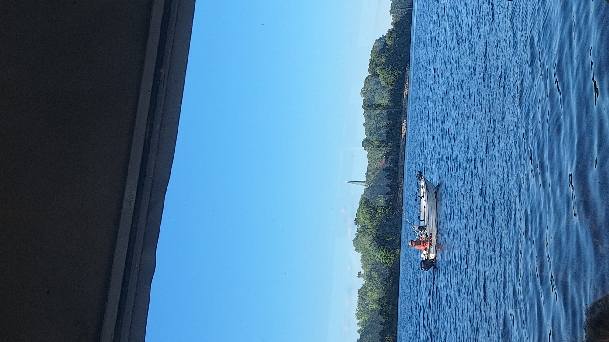 The image depicts a bright, clear day with a wide expanse of blue sky over a body of water. On the right side of the image, the water is a vibrant blue, reflecting the sky and showing gentle ripples. A small white boat with an outboard motor is visible on the water. A person wearing an orange life vest is seated in the boat, appearing to be engaged in an activity like fishing. The boat has "RANA" written on its side.
Along the left side of the image, a dark, textured surface dominates, suggesting a structure such as a bridge or overhang, casting a shadow. Beyond the water and the boat, a lush, green tree-covered shoreline extends into the distance. A slender steeple, likely belonging to a church, pierces the treeline, hinting at a settlement. The overall atmosphere is serene and peaceful, indicative of a summer day. The clear sky and bright sunlight suggest it is daytime.
The scene is set in Koknese, Latvia, as indicated by the context. No explicit emotions are conveyed, but the scene evokes a sense of tranquility and outdoor recreation.