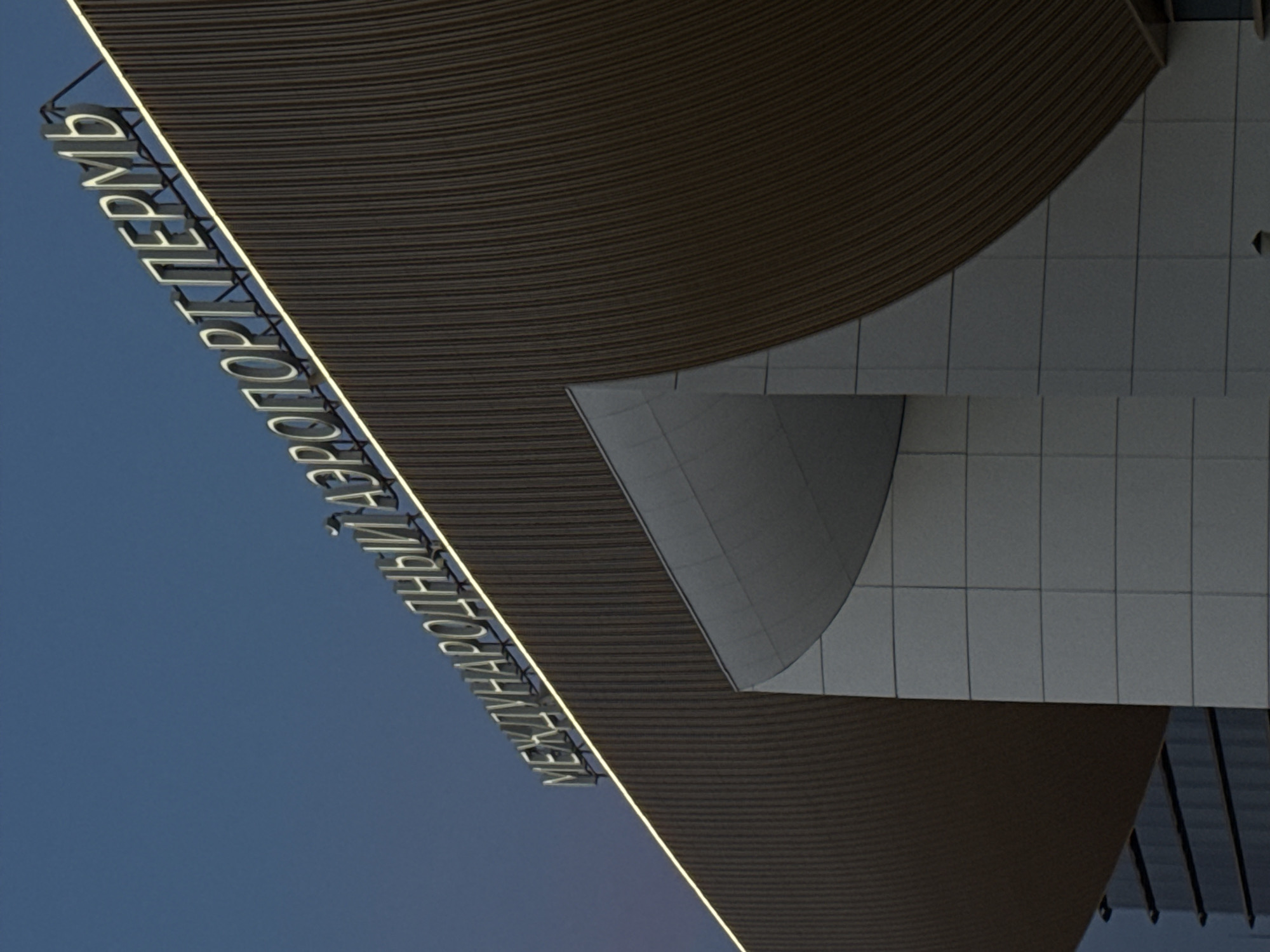 The image depicts the exterior of the Perm International Airport in Bolshoye Savino, Russia, at dusk or dawn, based on the blue sky. The architectural style features curved, horizontally ribbed roofing and facade elements. The name of the airport, "МЕЖДУНАРОДНЫЙ АЭРОПОРТ ПЕРМЬ," is illuminated in white letters on the edge of one of the roofs. The facade consists of rectangular tiles in a light gray color. The image is taken from a low angle, looking up at the structure.