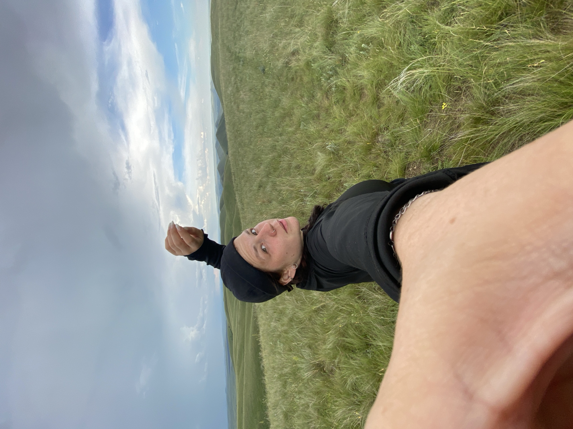 A young woman with brown hair, wearing a black beanie and a black sweatshirt, is taking a selfie outdoors on a grassy hill. Her left arm is extended towards the camera, with her hand blurred. She is looking at the camera with a neutral expression. The background shows rolling green hills under a cloudy sky with patches of blue. The scene appears to be in a rural, natural setting, possibly in or near Verkhneozernoe, Russia, given the landscape.