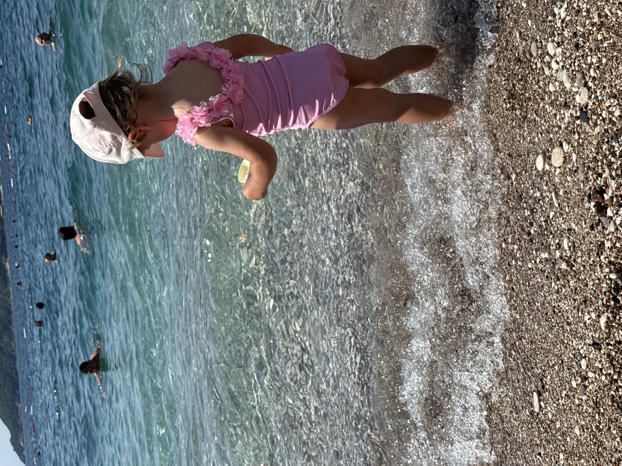 A young child wearing a pink swimsuit and a light pink cap stands in shallow water at a pebble beach in Bečići, Montenegro. The child is facing away from the camera and appears to be holding a yellow object in their right hand. The water is clear and turquoise, revealing the pebble seabed. In the background, several adults are visible swimming in the sea, and a tree-covered hillside forms the horizon. The sun is shining, casting reflections on the water.