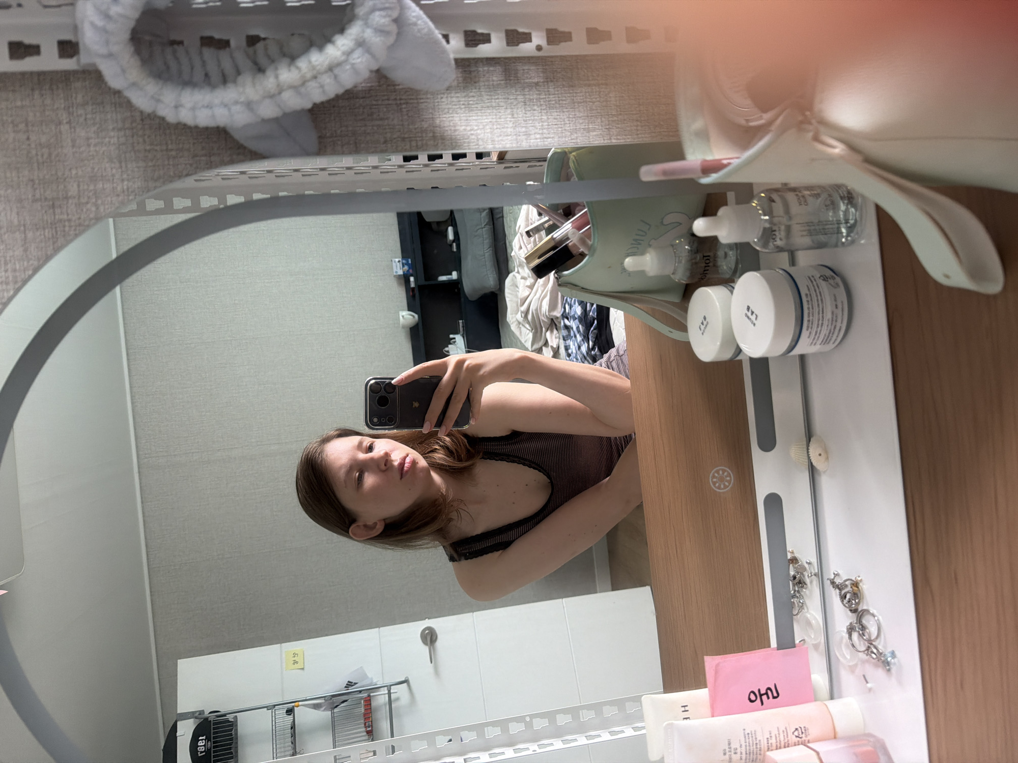 The image, taken in Dongducheon-si, South Korea, shows a young woman in her early 20s with light brown hair and fair skin, holding a smartphone to take a mirror selfie. She is wearing a dark, striped tank top. The background suggests a bedroom setting, with a bed partially visible behind her, covered in rumpled linens.
In the foreground, a vanity table is visible, featuring various cosmetic items. These include several bottles and jars of skincare products, a makeup bag filled with brushes and lipsticks, and a pink sticky note with Korean writing. Small decorative items like seashells and keychains are also scattered on the vanity. A large, oval mirror with a white frame reflects the scene. To the right of the mirror, a light blue, fuzzy headband with ears is attached to a white rack. The room's walls are textured, and a doorway with a silver handle is visible to the left. A metal shelving unit with various items is also present on the left side of the frame. The lighting appears to be natural, coming from an unseen source.