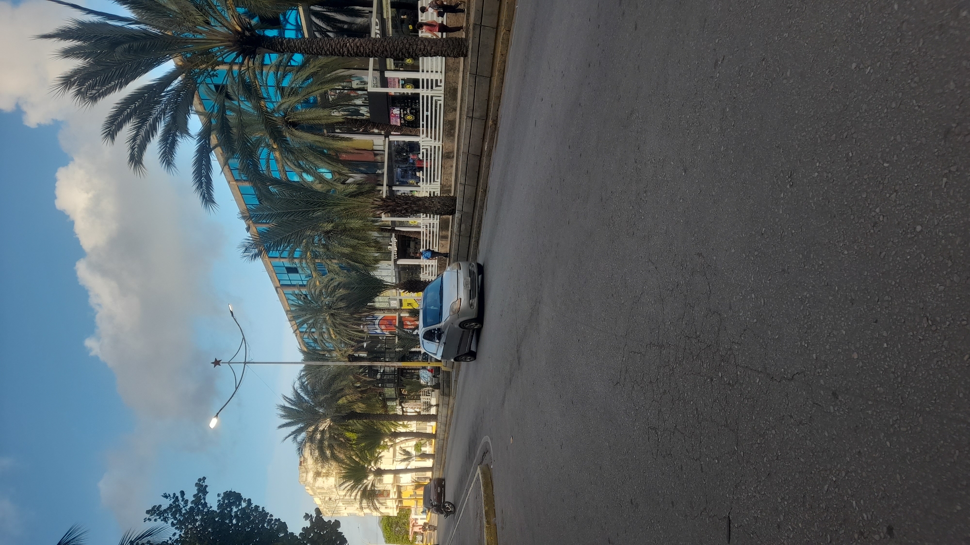 The image captures a street scene in Juan Griego, Venezuela, likely during the late afternoon or early evening, given the lighting and sky. A silver hatchback car is driving on the right side of the asphalt road, which shows some signs of wear and cracking. On the sidewalk, several palm trees are prominently displayed, framing a multi-story building with blue glass windows. The building appears to be commercial, with several storefronts visible. People are seen walking on the sidewalk in front of these establishments. Further down the street, a motorcycle is parked on the left side, and another vehicle is visible in the distance. A street lamp with a star-shaped ornament at its apex stands on the left side of the road. The sky is a clear blue with scattered white clouds.