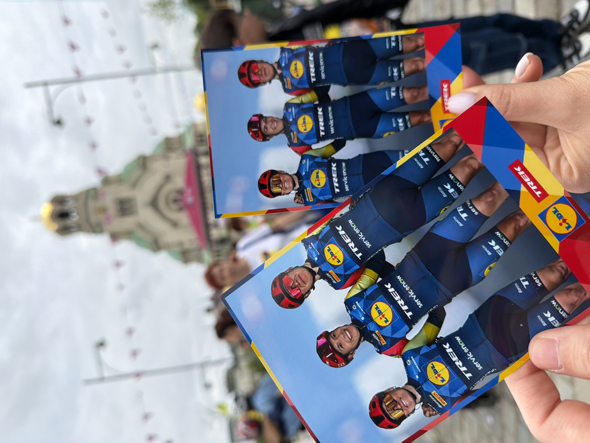 The image shows two rectangular photographs being held in a person's hand, with a blurred cityscape in the background. The photographs depict three female cyclists wearing matching blue, yellow, and red cycling jerseys, with the logos of Lidl and Trek visible on the jerseys. The background of the photographs appears to be a bright blue sky.
In the blurred background, the architecture of a city is discernible. The most prominent feature is a large, domed building with a golden roof. Based on the appearance of the domed building and its surroundings, it seems highly probable that the location is Sofia, Bulgaria, with the building likely being the Alexander Nevsky Cathedral. The sky is overcast, and there appear to be flags or decorations strung across the streets in the distance. There are people blurred out behind the photographs, which seem to suggest an active public space.