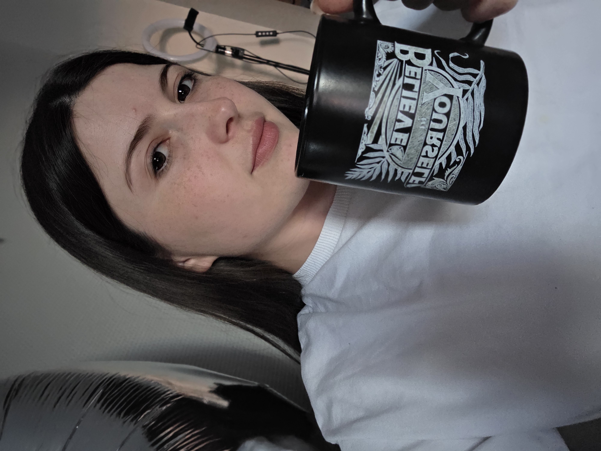 A young woman with dark hair and light skin is holding a black mug with a white design and text that reads "Believe in Yourself" in a mirror image. She is wearing a white long-sleeved shirt and has freckles on her face. The background is out of focus and appears to be an indoor setting with a large, shiny, dark object on the left, possibly a balloon, and some equipment with lights in the background on the right.