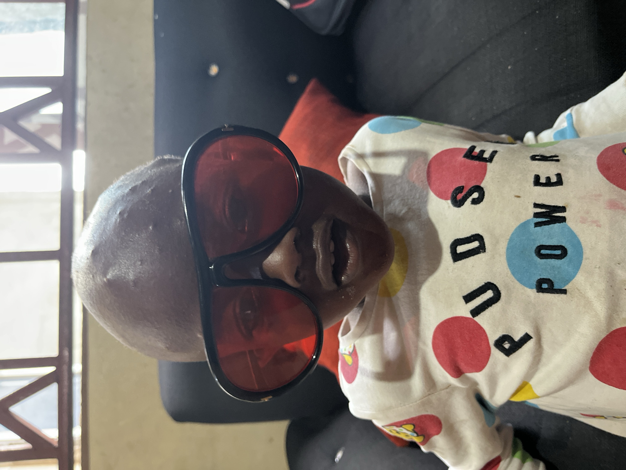 A baby is wearing oversized red sunglasses and a white shirt with red, blue, and yellow polka dots, and the words "PUDSE POWER" printed on it. The baby is on a black couch with a red pillow. A window with a grid pattern is visible in the background. The image was likely taken indoors. There is no information from the image itself to suggest the location is Badagry, Nigeria.