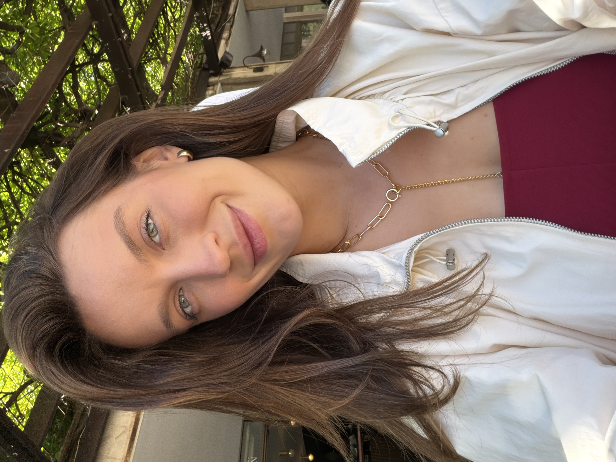 A young woman is featured in a close-up, slightly rotated selfie, with her head tilted towards the left. She has long, wavy brown hair, framing her face and cascading over her shoulder. Her green eyes are looking directly at the camera, and a soft, natural smile plays on her lips, adorned with a light pink tint. She wears a cream or off-white zippered jacket, partially open to reveal a burgundy top underneath. Around her neck, she sports a layered gold necklace, comprising a thicker paperclip chain and a delicate thinner chain. A small gold hoop earring is visible in her left ear.
The setting is outdoors during the daytime, characterized by soft, natural light. The background features abundant green foliage, possibly vines, growing over dark wooden beams, indicative of a pergola or similar garden structure. Glimpses of a building can be seen through the greenery. The woman appears relaxed and happy. No text is visible in the image. This scene is located in Budva, Montenegro.