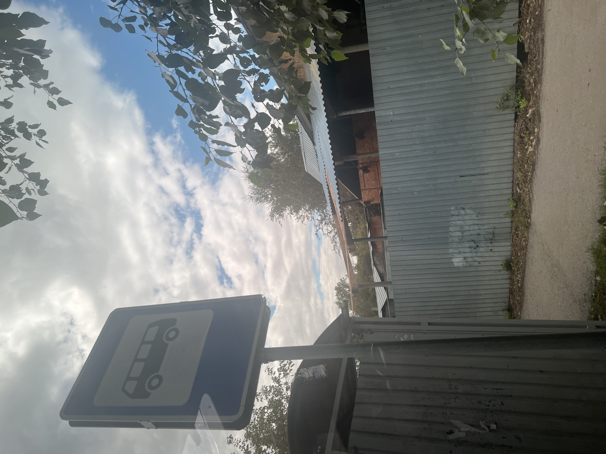 The image shows a bus stop sign in Ulyanovsk, Russia. The sign is blue and white and has a silhouette of a bus on it. The sign is mounted on a pole and is located next to a corrugated metal fence. Behind the fence, there are some buildings and trees. The sky is cloudy and blue. There is a road in the foreground. The overall scene appears to be an outdoor environment in a semi-urban or rural setting.