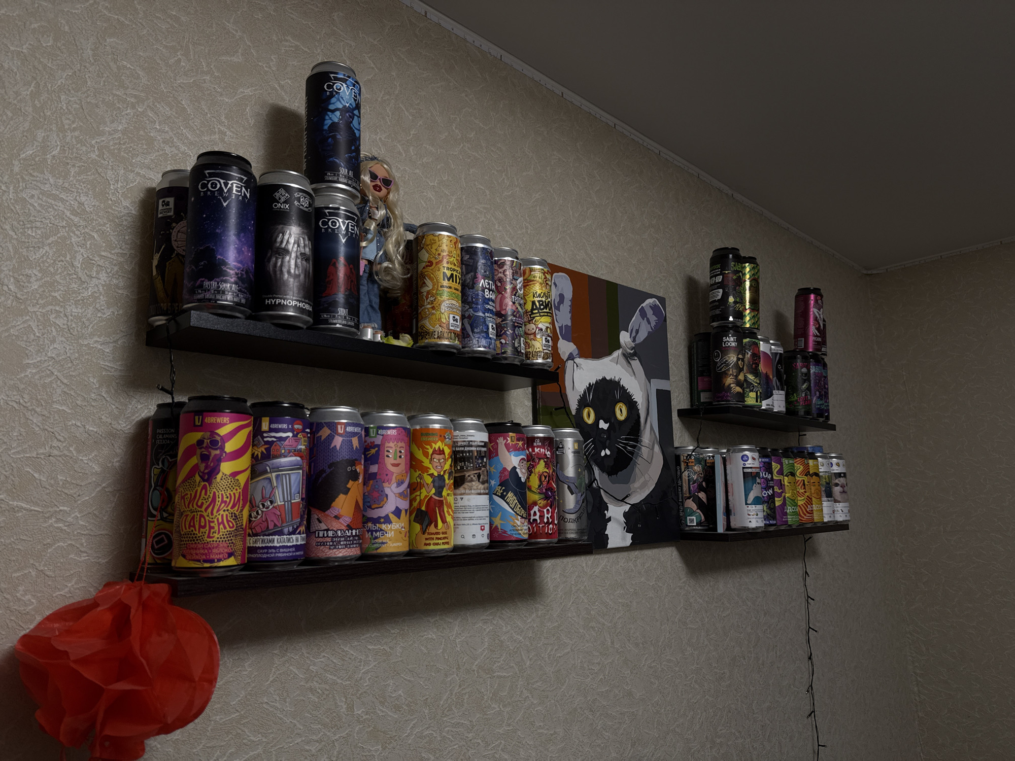 The image shows shelves of canned beverages, likely beer or cider, in a domestic setting. The shelves are dark, narrow, and mounted on a textured, light-colored wall. On the top shelf, from left to right, are several cans with intricate designs, including one labeled "COVEN BREWERY" with a dark blue and purple swirling sky, and another with a dark, gothic-style image of eyes. A doll with blonde hair and sunglasses is also present on this shelf. Further to the right, more cans with colorful designs are visible. The bottom shelf is also filled with cans, many featuring cartoonish illustrations and bold text in Russian. A red paper lantern hangs from the left side of the bottom shelf. A string of small, dark-colored lights is visible on the wall behind the bottom shelf, extending downwards. A large framed artwork or print featuring a stylized black cat with yellow eyes is mounted on the wall above the bottom shelf, partially obscured by cans. The lighting in the scene is dim, creating shadows. The overall environment suggests a collection or display of craft beverages in a home.