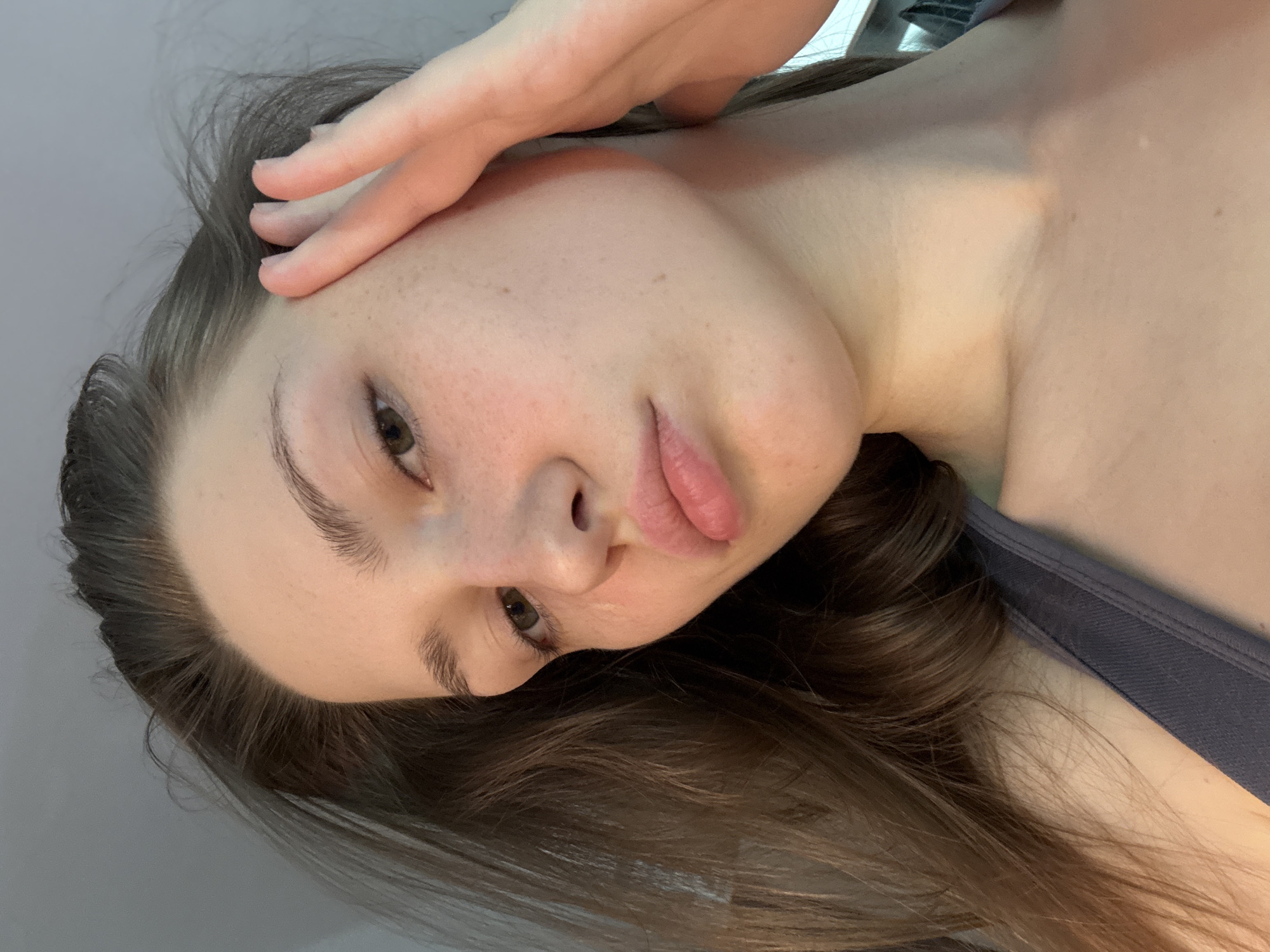 The image displays a close-up portrait of a young woman, captured from an upward angle. The woman's head is tilted to the left, suggesting she might be lying down or leaning back. She has fair skin, adorned with numerous subtle freckles visible across her face, forehead, and upper chest. Her eyes are brown, and her full lips are slightly parted in a soft pout. Her eyebrows are natural and well-defined. Her long, wavy hair, a rich dark brown, cascades around her head and shoulders. Her right hand is gently placed near her temple and hairline, with her fingers slightly spread. She is wearing a dark blue or grey strap of clothing, likely a tank top or camisole, visible on her right shoulder and upper chest. The background is a plain, light grey or off-white surface, indicating an indoor setting. The lighting is soft and diffused, creating a gentle illumination without harsh shadows. Her expression appears contemplative or perhaps a little melancholic. No text is visible within the image. This portrait is situated in Saint Petersburg, Russia.