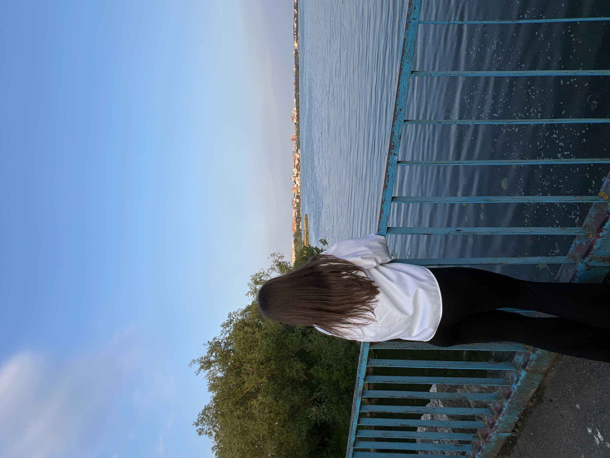 The image shows a person standing on what appears to be a bridge or pier, looking out over a large body of water. The person is seen from behind, leaning against a blue metal railing. They have long, dark brown hair, wear a white long-sleeved shirt, and black pants.
The scene is dominated by the expansive blue water on the right, which shows ripples on its surface, reflecting the sky. On the far bank of the water, a distant city or town skyline with numerous buildings is visible under a clear, light blue sky. To the left of the person and below the skyline, lush green trees are visible, extending along the bank.
The railing the person is leaning on is painted blue, but shows signs of wear and rust, suggesting it might be an older structure. The sky above is mostly clear with a subtle gradient from a deeper blue to a lighter hue towards the horizon. The lighting suggests late afternoon or early evening, with soft, ambient light illuminating the scene. The overall impression is one of peaceful observation or contemplation of the scenic landscape. There is no visible text in the image. Given the provided context, the location is Ternopil, Ukraine, likely overlooking Ternopil Lake.
