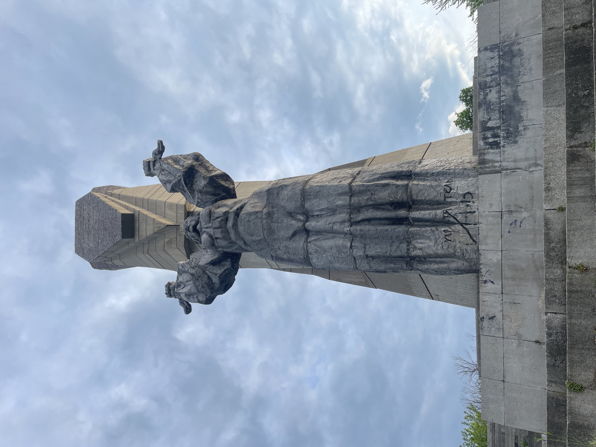 The image shows the Defenders of Stara Zagora Memorial Complex in Stara Zagora, Bulgaria. The monument features a large stone statue of a figure on top of a concrete structure, set against a cloudy sky. There are no people visible in the image. The base of the concrete structure has some graffiti.