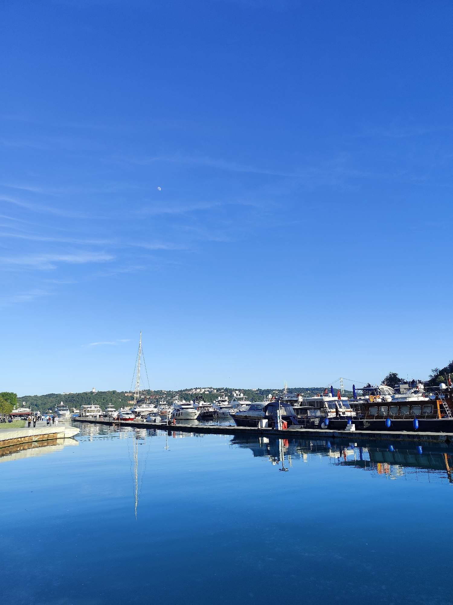 The image depicts a marina scene in Sarıyer, Turkey. A clear blue sky, with a faint moon visible, stretches above a wide expanse of water. Multiple yachts and boats are docked along a concrete pier. Their reflections are visible in the calm water. The land behind the marina features lush green trees and residential buildings. In the far left, a few people are walking along a concrete path. A single sailboat mast extends high above the other boats.