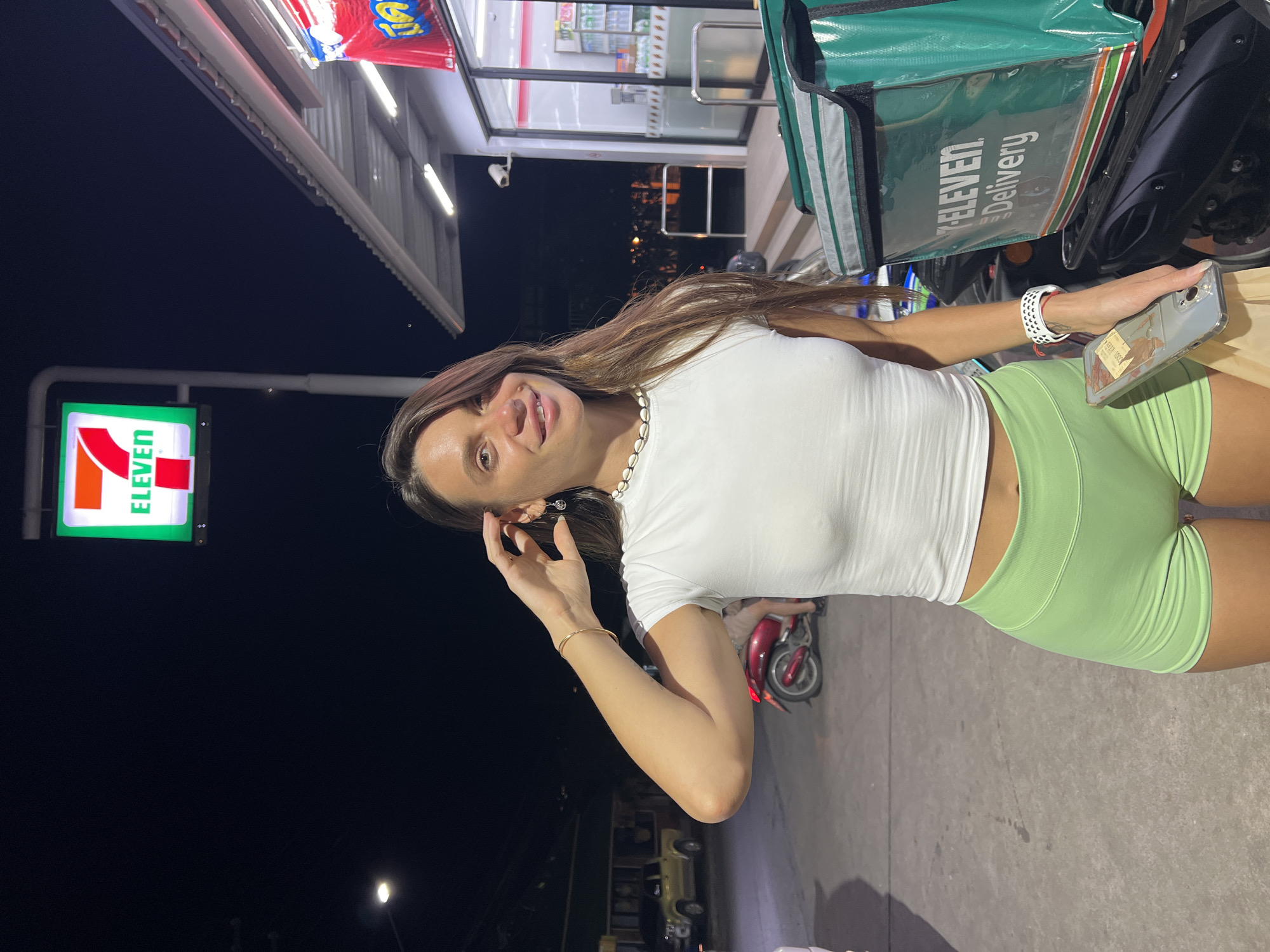 A young woman with long, brown hair stands in front of a 7-Eleven convenience store at night. She is wearing a white t-shirt and light green shorts. She is holding a smartphone in her right hand and her left hand is near her face. The 7-Eleven store has a prominent illuminated sign. A delivery scooter with a green insulated bag is parked next to the woman. The background shows the interior of the store with shelves stocked with products. The ground is paved, and a street light is visible in the distance. The time of day is night, indicated by the dark sky and artificial lighting. The setting appears to be in Thailand, as indicated by the location context. There is no indication of the weather. The woman's expression appears neutral or slightly playful. Visible text includes "7-ELEVEN Delivery" on the scooter bag and "7-ELEVEN" on the store sign.