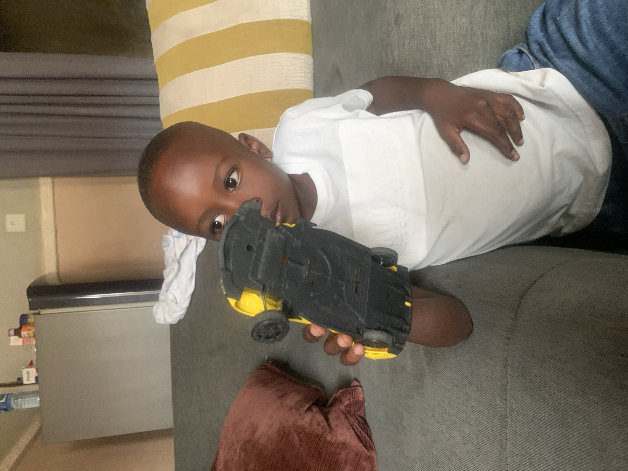 A young Black boy, appearing to be around 3-4 years old, sits on a gray couch in what is presumed to be a living room in Sabon Tasha, Nigeria. He is wearing a white t-shirt and blue jeans. He holds a yellow and black toy car upside down in his right hand, examining its underside. The boy is looking off to his right. To his left, a dark red pillow rests on the couch. Behind him, a yellow and white striped pillow is visible. In the background, a refrigerator and some items on top of it can be seen. The overall environment suggests a casual, indoor setting.