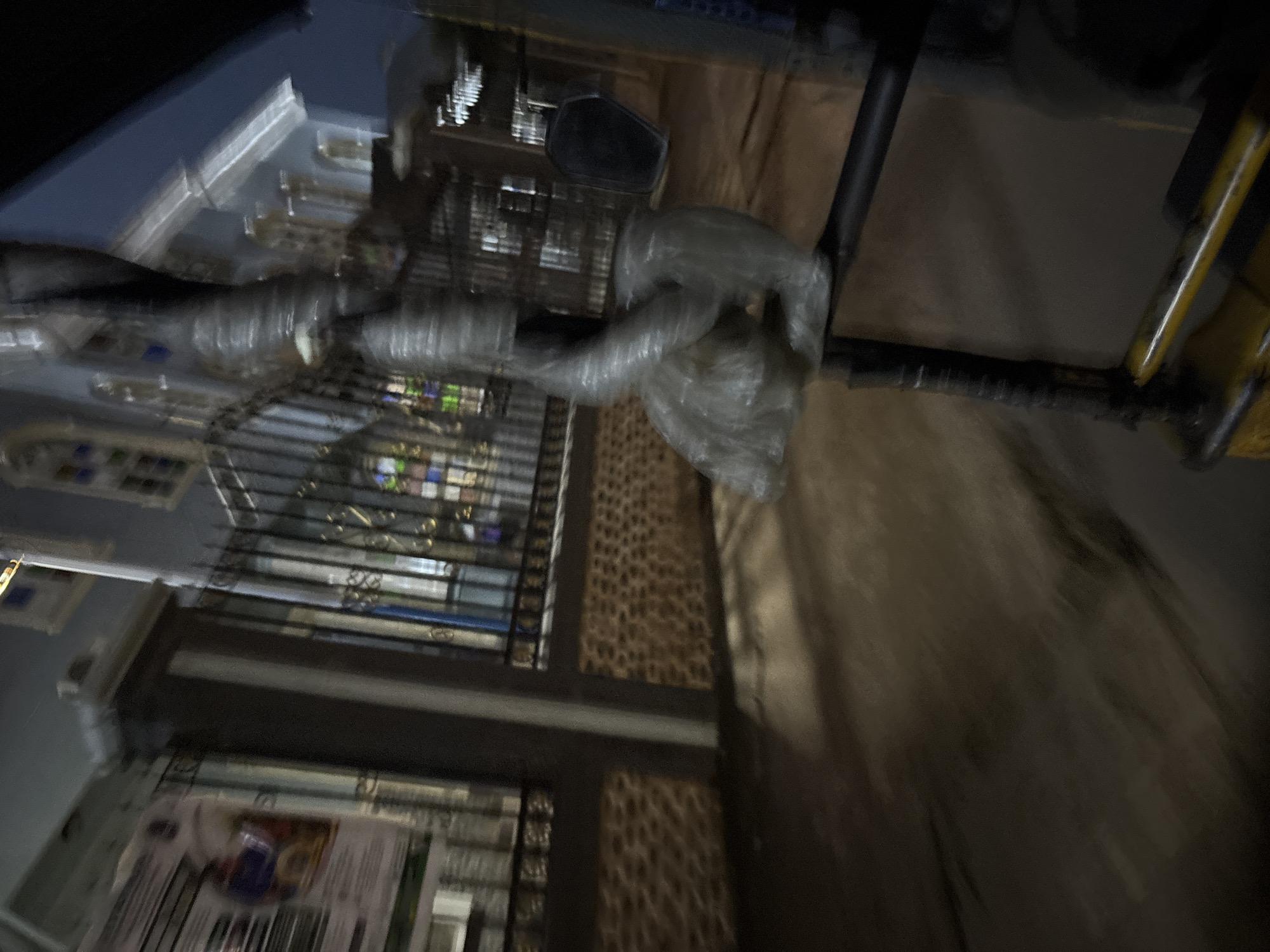 The image depicts a blurry night scene in front of a building with a black metal fence. The building's facade appears to have stained-glass windows, suggesting it could be a church or a similar institution. The fence is ornamental, with vertical bars and decorative elements at the top and bottom. To the left of the fence, a poster is partially visible, displaying an image of a person in a blue garment. The foreground features a dark, paved surface, likely a road or sidewalk, with some indistinct shadows. In the mid-ground, a plastic-wrapped object, possibly a statue or sculpture, is precariously perched on a black pole. The overall lighting suggests artificial illumination from streetlights or interior building lights. The motion blur indicates that the photograph was taken from a moving vehicle. The image is identified as being taken in Onitsha, Nigeria.