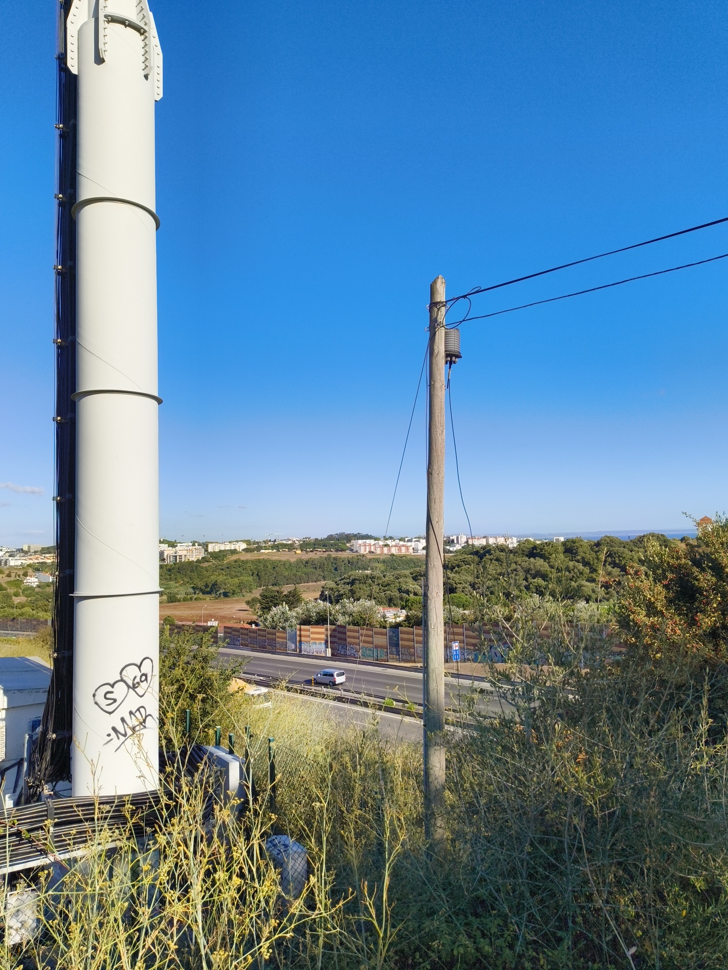 The image shows a telecommunications tower on the left, with graffiti consisting of two hearts and the markings "S 69" and "-MAB" visible on its surface. A wooden utility pole with electrical wires stretches across the right side of the frame. In the background, a highway runs through a landscape dotted with trees and buildings, suggesting a semi-urban or suburban environment. The scene is set under a clear blue sky. The overall impression is one of a functional, everyday outdoor setting. While the exact location within Cascais, Portugal, is not explicitly shown, the presence of such infrastructure and landscape is common in the region.