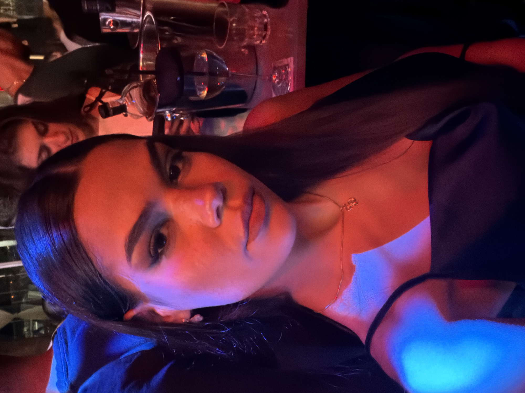 An indoor scene, likely a bar or nightclub in Varna, Bulgaria, is depicted at night, characterized by dim, colored ambient lighting. The central subject is a young woman with dark hair, shown from the chest up, looking directly at the viewer with a neutral expression. Her face and upper chest are illuminated by a strong red light, while a blue light falls across her neck and shoulder area, creating a dramatic, high-contrast effect. She is wearing a dark top and a delicate necklace with a small pendant. In the blurred background, partially visible on the left, is another person, possibly a woman, with long dark hair, along with various glasses and barware on a surface. The image's composition, with the subject oriented horizontally, emphasizes the vibrant and atmospheric lighting of the venue. No text is visible.