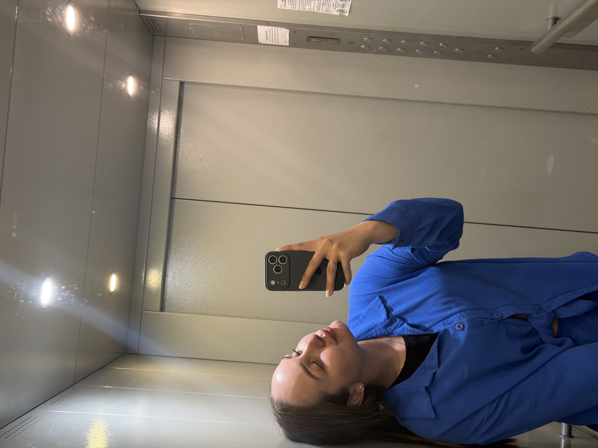 A woman is taking a selfie in what appears to be an elevator. She is wearing a blue shirt and holding a black smartphone. The elevator has a white interior with recessed lights and a panel with buttons, suggesting it serves multiple floors. The image does not provide enough information to determine the specific location in the city of Chaikovsky, Russia.