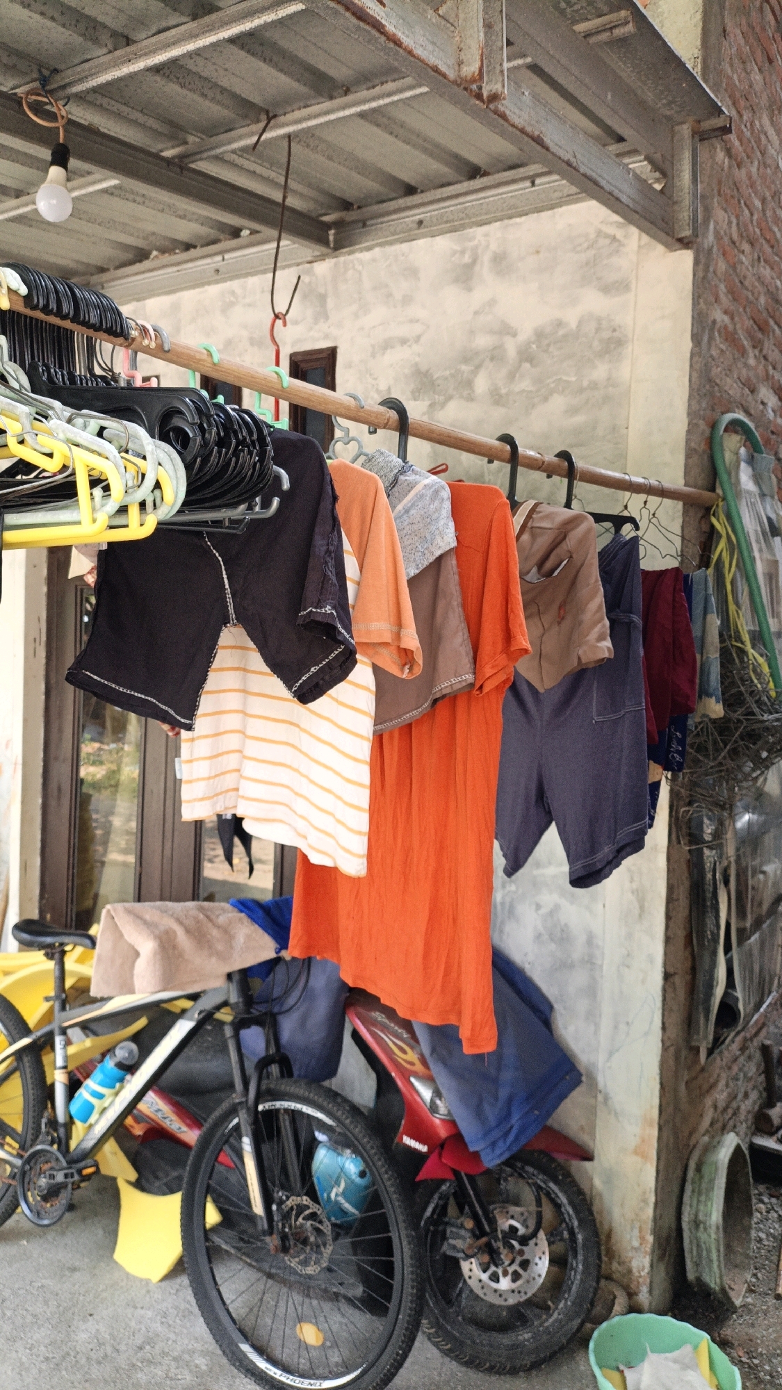 This is an outdoor image taken in Cikal, Indonesia, showing a clothesline filled with various items of clothing hung on hangers. The clothes are predominantly in shades of orange, brown, and blue, with a few striped items. Beneath the clothesline, a bicycle and a red motorcycle are visible, partially obscured by the hanging garments. The background consists of a textured wall and a corrugated metal roof supported by metal beams. The overall scene suggests a residential or utilitarian outdoor space.