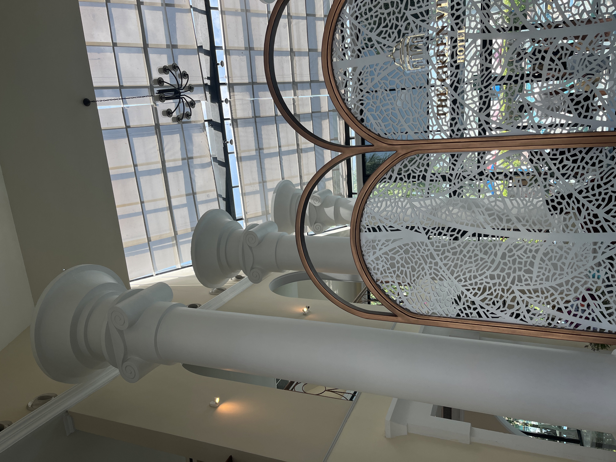 The image captures the interior of a building, likely a hotel lobby, featuring an atrium with a large glass roof. Prominently visible are several tall, white Corinthian columns that extend upwards towards the ceiling. In the foreground, a decorative screen with an intricate white pattern, possibly representing branches or foliage, and a bronze frame divides the space. Through the screen, the words "THE GREEN VIEW HOTEL" are discernible, suggesting the location. A multi-armed chandelier hangs from the ceiling. The overall ambiance suggests a well-lit, spacious interior with classical architectural elements. The image does not show any people. The location is identified as Bang Sao Thong Subdistrict, Thailand.
