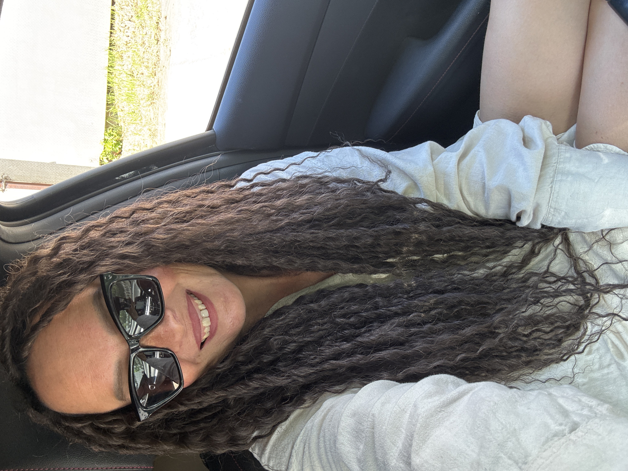 A woman wearing sunglasses and a light-colored shirt is sitting in a car. Part of her legs are visible. The location is inside a car; outside the window appears to be an exterior setting with grass and a white wall. The location within the city of Chaikovsky, Russia cannot be determined from the visual information provided.