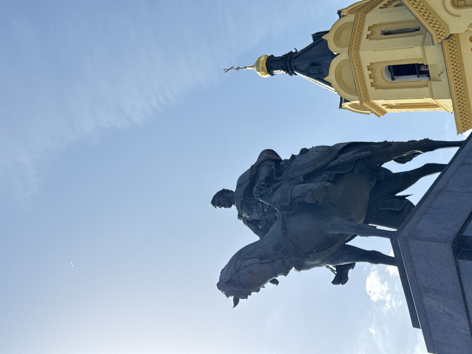 The image shows a statue of a man on horseback with a church tower in the background. The statue is bronze and appears to be atop a gray stone pedestal. The background consists of a blue sky with a few faint clouds and part of a yellow and black church tower with a golden dome and cross. The location is likely Nizhny Novgorod, Russia, specifically the Minin and Pozharsky Square, where a statue of Kuzma Minin and Dmitry Pozharsky stands with the Archangel Michael Cathedral nearby.