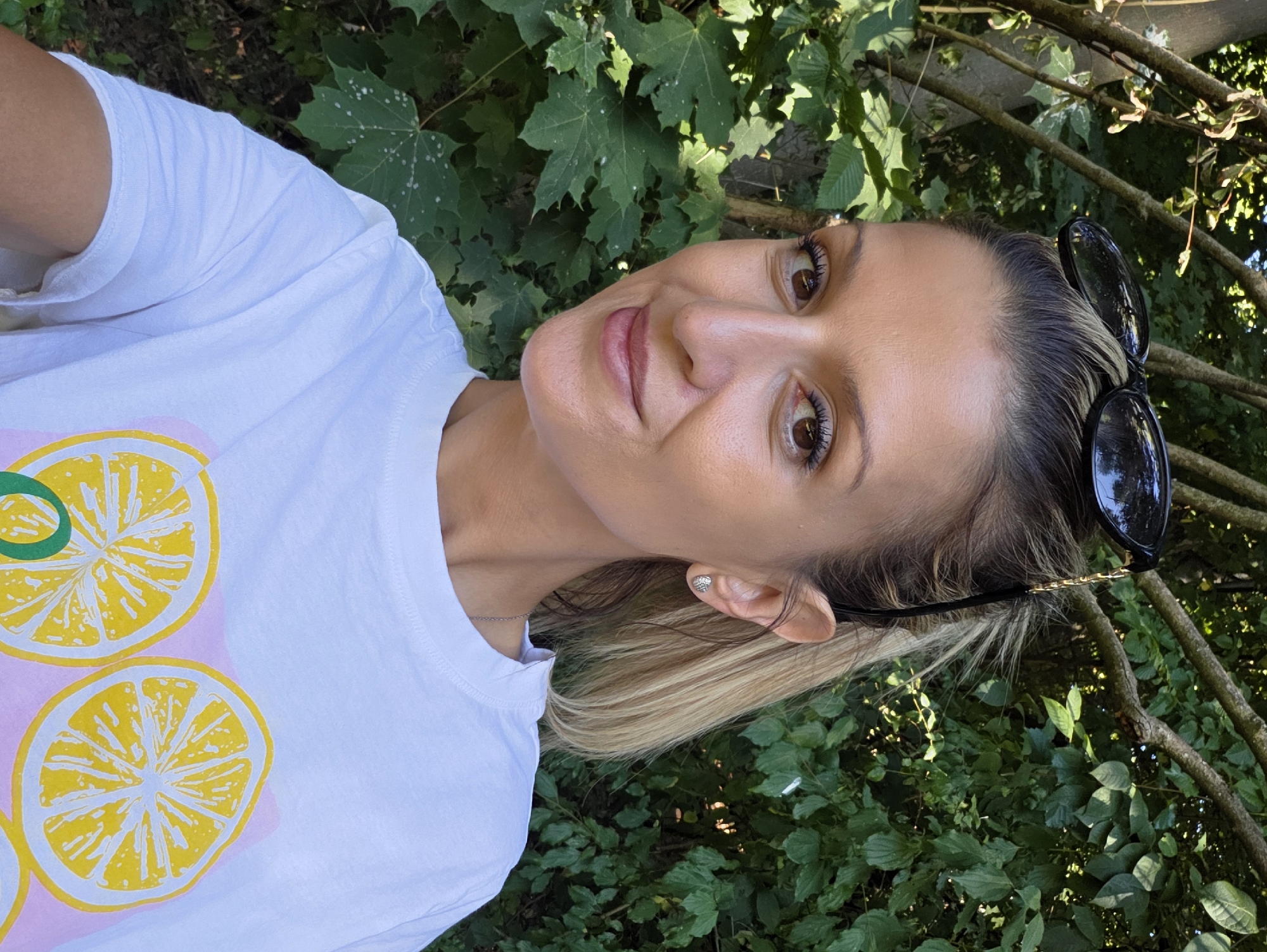A woman with blonde hair pulled back in a ponytail and sunglasses perched on her head smiles at the camera. She is wearing a white t-shirt with a graphic print of lemon slices. She is outdoors, with lush green foliage behind her. The image appears to be a selfie taken during the daytime. The woman is wearing subtle makeup and a small silver earring. Her t-shirt has a faint pattern of what appears to be more fruit slices.