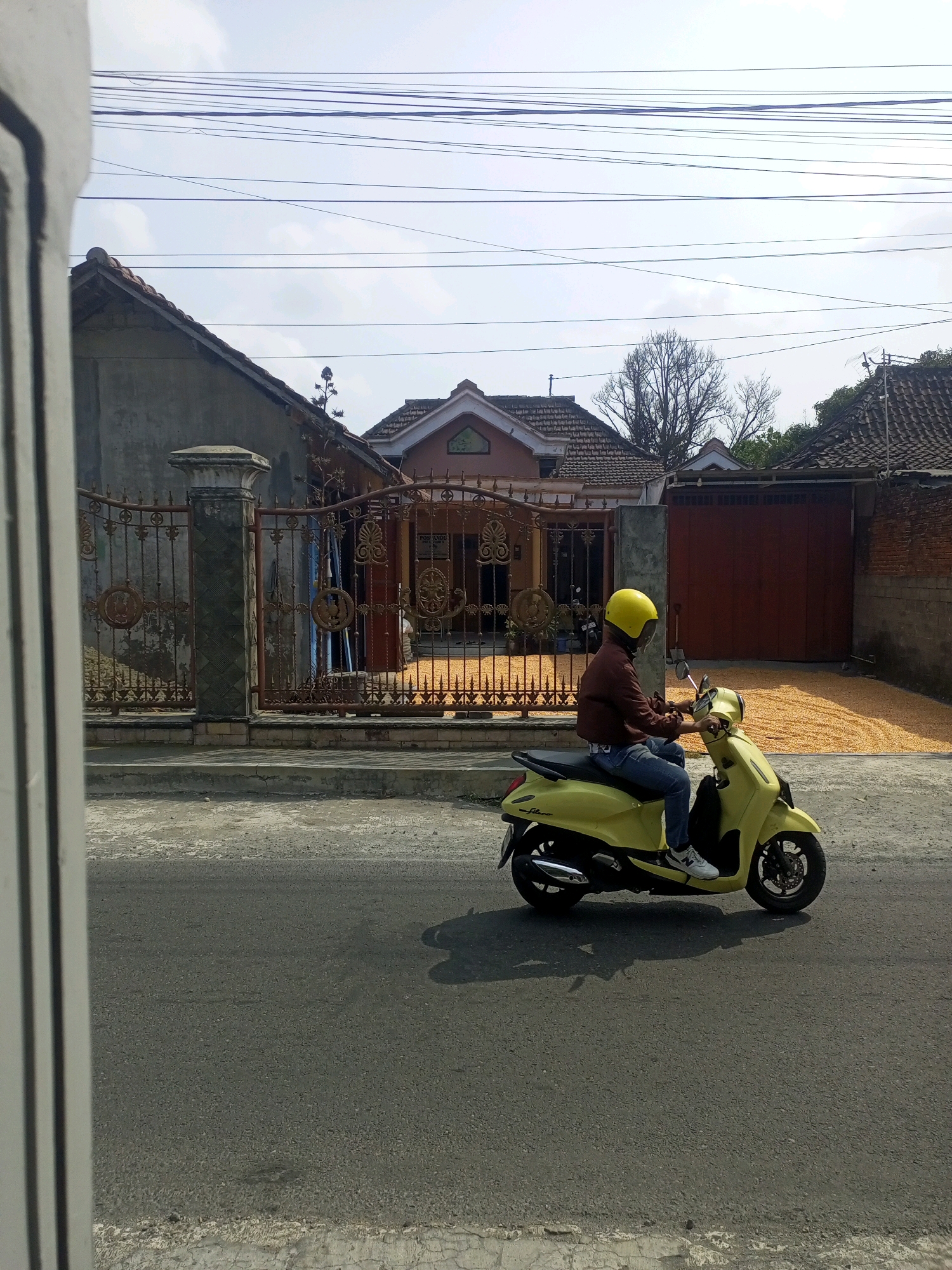 A person wearing a yellow helmet, a brown jacket, and blue jeans rides a yellow scooter on an asphalt road. The scooter is positioned in the right half of the image, facing left. In the background, a house with a red-tiled roof and an ornate metal fence are visible. A large pile of dried corn kernels is spread out on the ground in front of a closed red garage door. Overhead, multiple electrical wires crisscross the sky. The scene appears to be in the city of Ngasem, Indonesia, given the architectural style and the presence of drying corn.