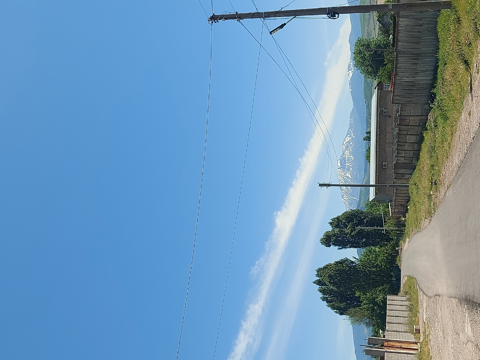 The image depicts a view in Tavaksay, Uzbekistan. A narrow asphalt road curves slightly to the right. Parallel to the road, there's a mix of vegetation, including grass and trees. A long, windowless building with a weathered wooden fence sits adjacent to the vegetation. In the background, snow-capped mountains are visible. An electric pole with wires dominates the left side of the frame, reaching into a clear blue sky with a wispy cloud formation. The light suggests it's daytime.