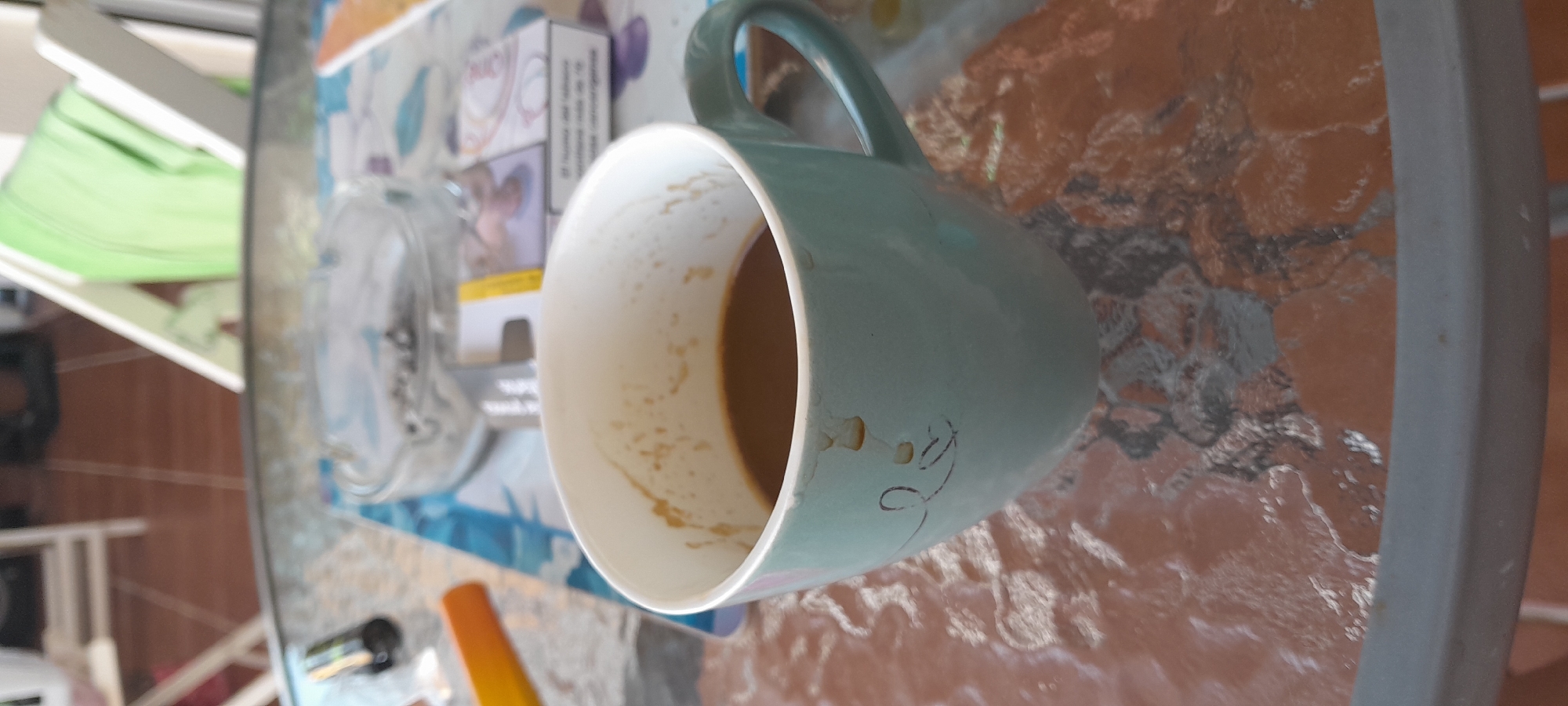 The image presents a close-up, elevated view of a light teal ceramic mug, partially filled with dark brown liquid, likely coffee. Visible coffee residue adheres to the interior rim and exterior surface of the mug, which also features a faint, simple linear drawing on its side. The mug rests on a textured glass tabletop exhibiting an underlying brown-orange hue, with a gray table rim visible in the foreground. Directly behind the mug, on the table, are a clear glass ashtray containing multiple cigarette butts and ash, and a partially obscured white cigarette pack with a yellow stripe. Textual elements on the cigarette pack, "¡FUMAR MATA!" (Smoking Kills!) and "PRODUCE CANCER" (Causes Cancer), indicate its origin in a Spanish-speaking region. To the left of the ashtray, a blue and white patterned object is visible, alongside an orange and yellow item and a dark cylindrical object. The background is blurred, showing a tiled floor composed of brown squares with white grout lines, segments of white chairs (one draped with a green fabric), and a light-colored wall or doorframe. No individuals are present in the scene, which is located in Manilva, Spain.