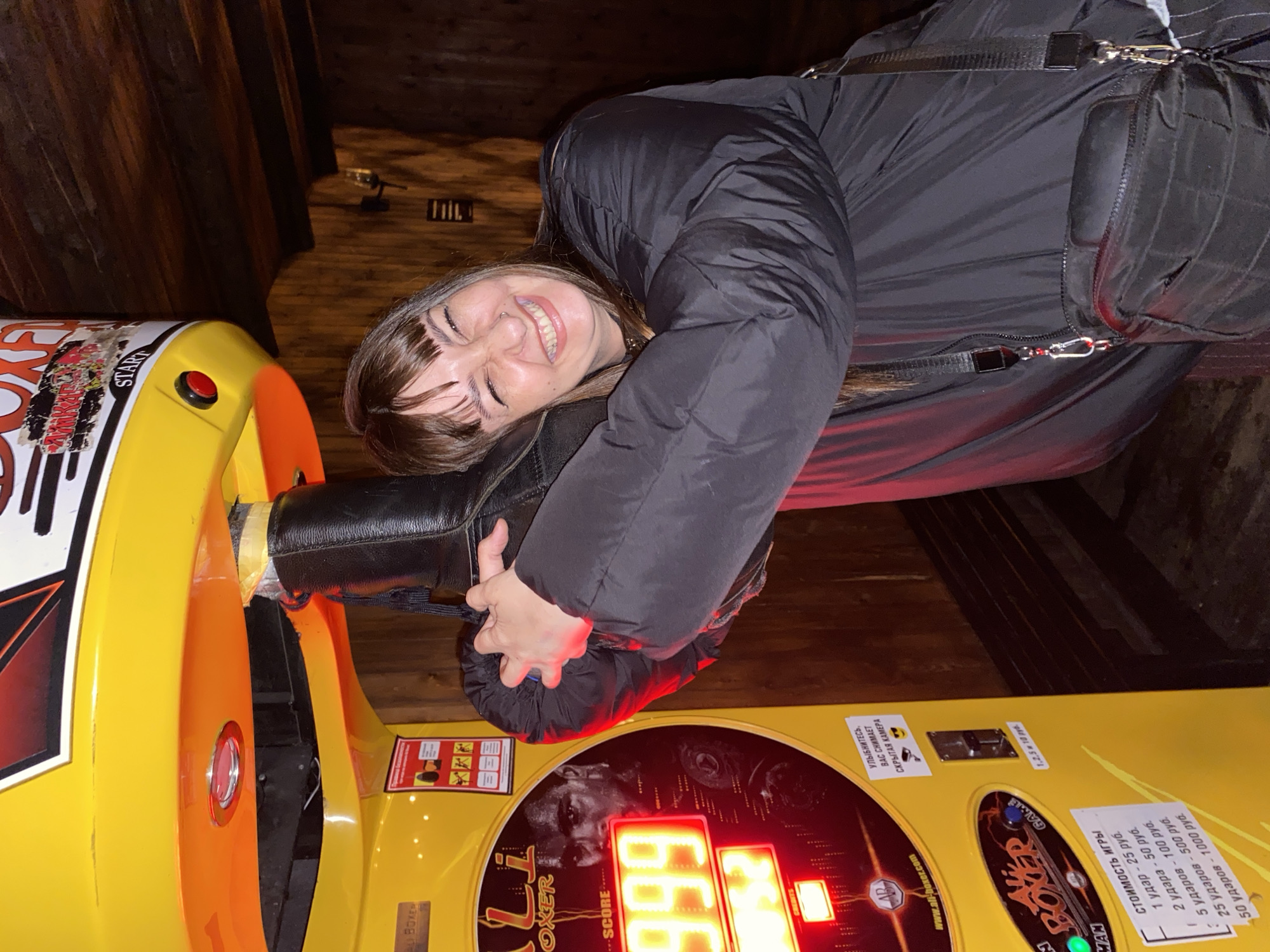 An image captures a young woman joyfully interacting with a yellow and orange arcade boxing machine in an indoor setting, likely an entertainment venue in Votkinsk, Russia. The scene is dimly lit, suggesting evening or nighttime, with dark wooden paneling visible in the background.
The woman, with long brown hair and bangs, is wearing a black puffer jacket and a black shoulder bag. She is leaning against the boxing machine, embracing part of it, and smiling broadly with her eyes mostly closed, conveying a sense of amusement and happiness.
The prominent arcade machine is branded "BOXER" and "Li BOXER". Its digital display shows a score of "666". An image resembling boxer Mike Tyson is visible on the machine's console. Various text elements in Russian are visible: "СТОИМОСТЬ ИГРЫ" (Game Cost) details prices for hits (1 hit - 25 rubles, 2 hits - 50 rubles, 5 hits - 100 rubles, 25 hits - 500 rubles, 50 hits - 1000 rubles). The coin slot indicates acceptance of "1,2,5 и 10 руб." (1, 2, 5, and 10 rubles). Another sign playfully warns, "УЛЫБНИТЕСЬ, ВАС СНИМАЕТ СКРЫТАЯ КАМЕРА" (Smile, a hidden camera is watching you). The machine also features "START" and the website "www.all-boxer.com". The overall impression is one of fun and leisure at an arcade.