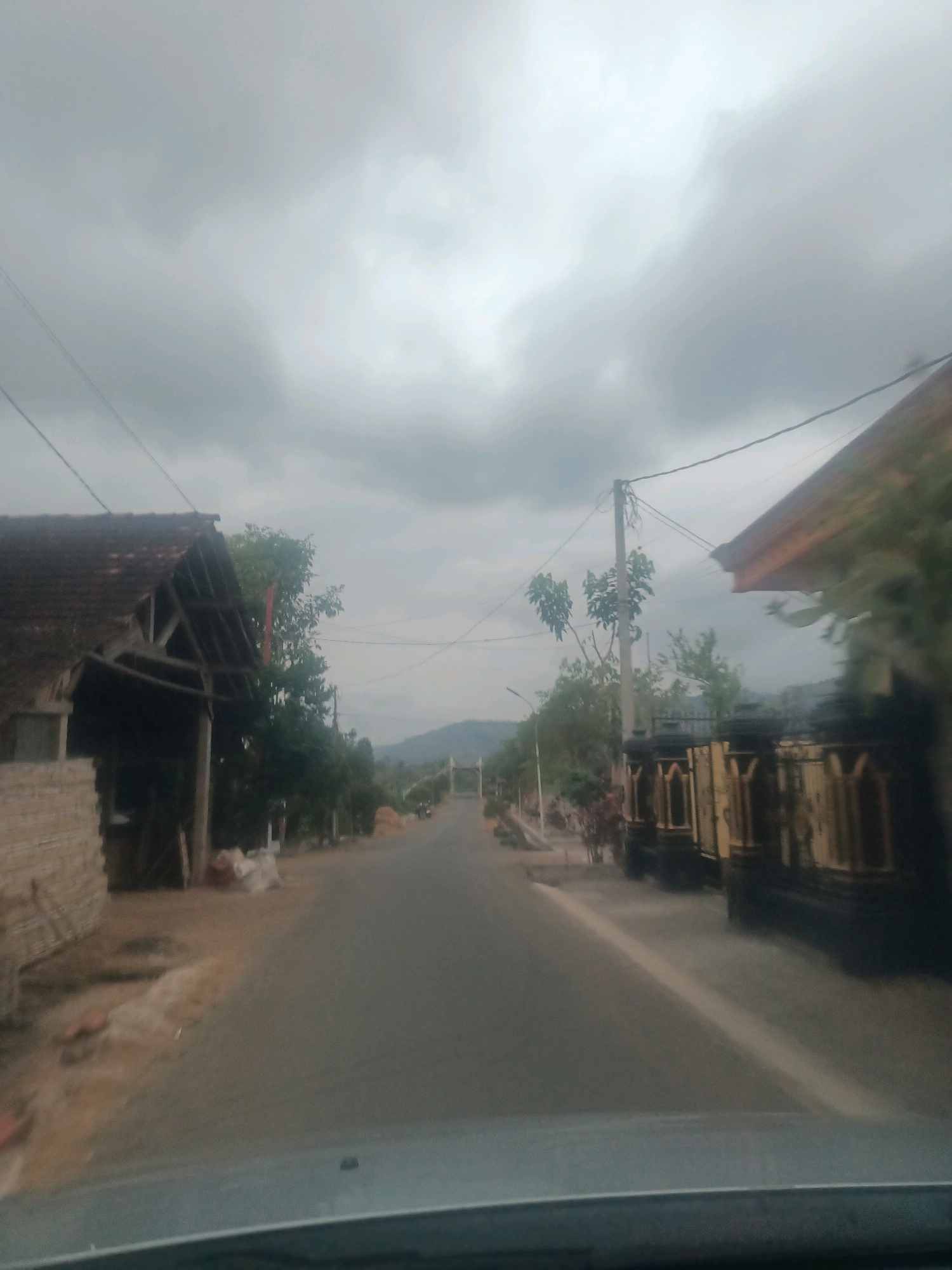 The image depicts a slightly blurred view from inside a vehicle looking out onto a paved road in a rural area, likely Gandusari, Indonesia. The sky is overcast with thick grey clouds, creating a subdued light. On the left side of the road, there is a dilapidated structure with a tiled roof and exposed wooden beams. Adjacent to it, a wall constructed of bricks is visible. Further down the road on the left, trees and vegetation line the path. On the right side of the road, a fence with decorative pillars and arches can be seen, with a portion of a building's roof extending into the frame. The road itself is empty, with a single white line marking the center. In the distance, hills are visible under the cloudy sky. The overall impression is that of a quiet, possibly overcast day in a village setting.
