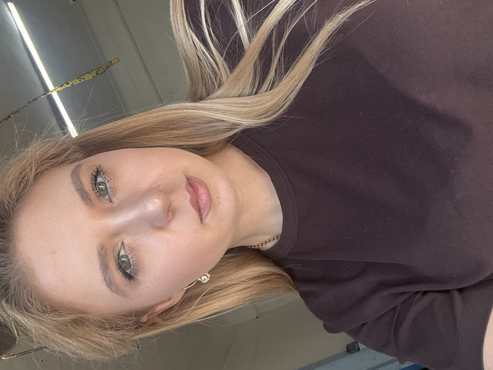 A blonde woman is shown from the chest up, looking into the camera. She has light skin and is wearing makeup, including mascara and lip gloss. She has on a dark brown crew neck shirt and a gold chain necklace. A gold earring with a clear stone is visible on her left ear. Her blonde hair is long and wavy. In the background, a fluorescent light fixture is visible on the ceiling, and a yellow object with black spots hangs from it. The environment appears to be indoors, possibly a garage or workshop, with a concrete floor and a light-colored wall.