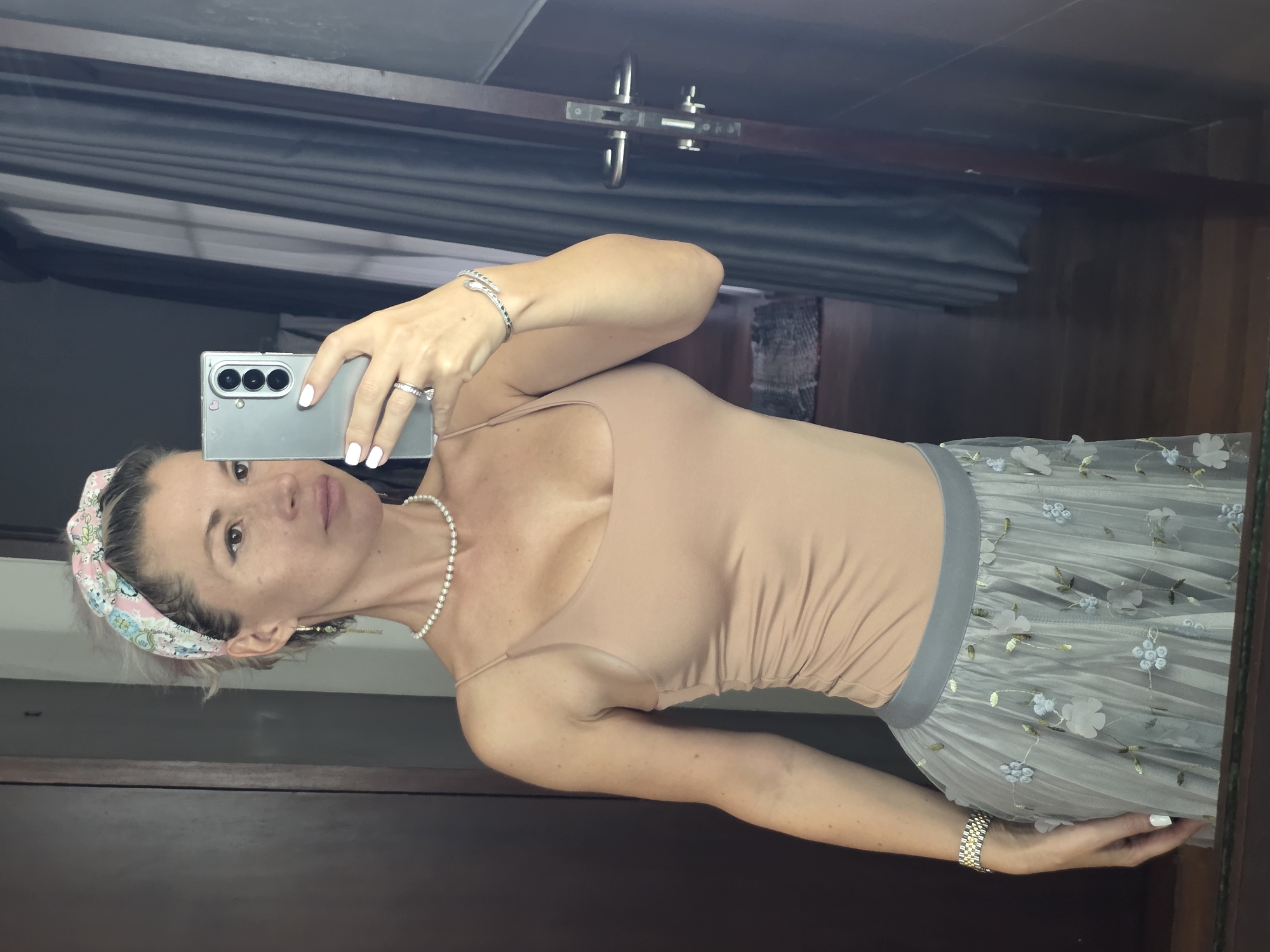 An eye-level, medium shot captures a woman taking a selfie in a mirror. The woman is standing in what appears to be a hotel room in North Kuta, Indonesia. She is wearing a beige spaghetti strap top, a sheer grey skirt adorned with floral appliques, a pearl necklace, a headband, and multiple bracelets and rings. Her nails are painted white, and she is holding a smartphone in her right hand, which is angled to capture her reflection. The background includes a wooden door frame, dark grey curtains, and a wooden floor.