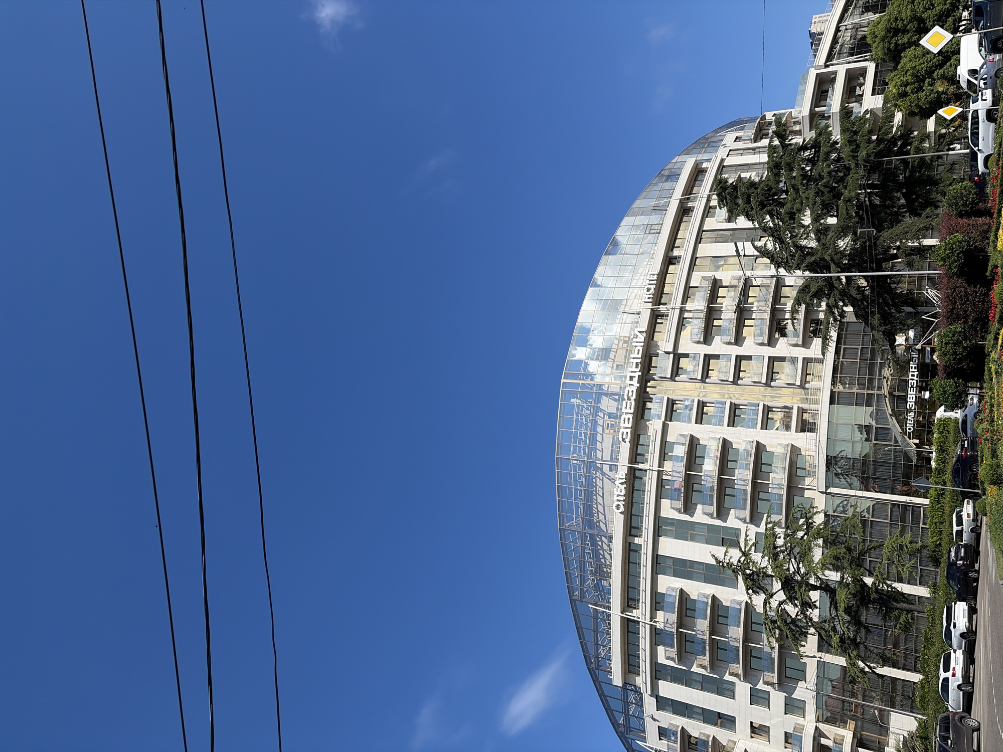 This is a daytime, eye-level, medium shot of the curved facade of a hotel building, with the clear blue sky dominating the upper portion of the frame. Three dark power lines are visible horizontally across the top of the sky. The hotel, identified by white lettering on its glass-domed roof that reads "ОТЕЛЬ ЗВЕЗДНЫЙ" (Hotel Zvezdny), is a multi-story structure with a modern, semicircular design. The building features numerous balconies with glass railings and rows of windows. Some of the windows reflect the sky and clouds. In front of the hotel, there is a street with several cars parked and driving. Green trees, including a tall evergreen, are planted in front of the building, and there are flower beds and bushes along the sidewalk. The scene appears to be in a city, likely Sochi, Russia, given the hotel's name and architectural style which are consistent with resorts in the area.