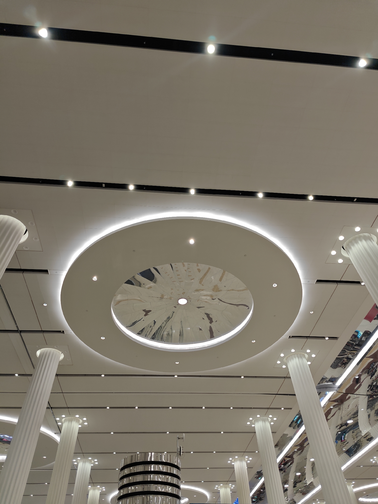 The image depicts the interior of a modern architectural space, likely a shopping mall or public building in Dubai, United Arab Emirates. The ceiling is a prominent feature, with a large circular recessed area illuminated by a ring of soft white light. At the center of this circle, a reflective surface with a marbled pattern is visible. Several tall, fluted white columns extend upwards towards the ceiling, some with smaller circular light fixtures at their tops. The ceiling is further adorned with a linear arrangement of recessed lighting. In the upper portion of the image, a long, dark horizontal element runs across the width of the frame, punctuated by several small, bright lights. The overall impression is one of spaciousness, modern design, and sophisticated lighting.