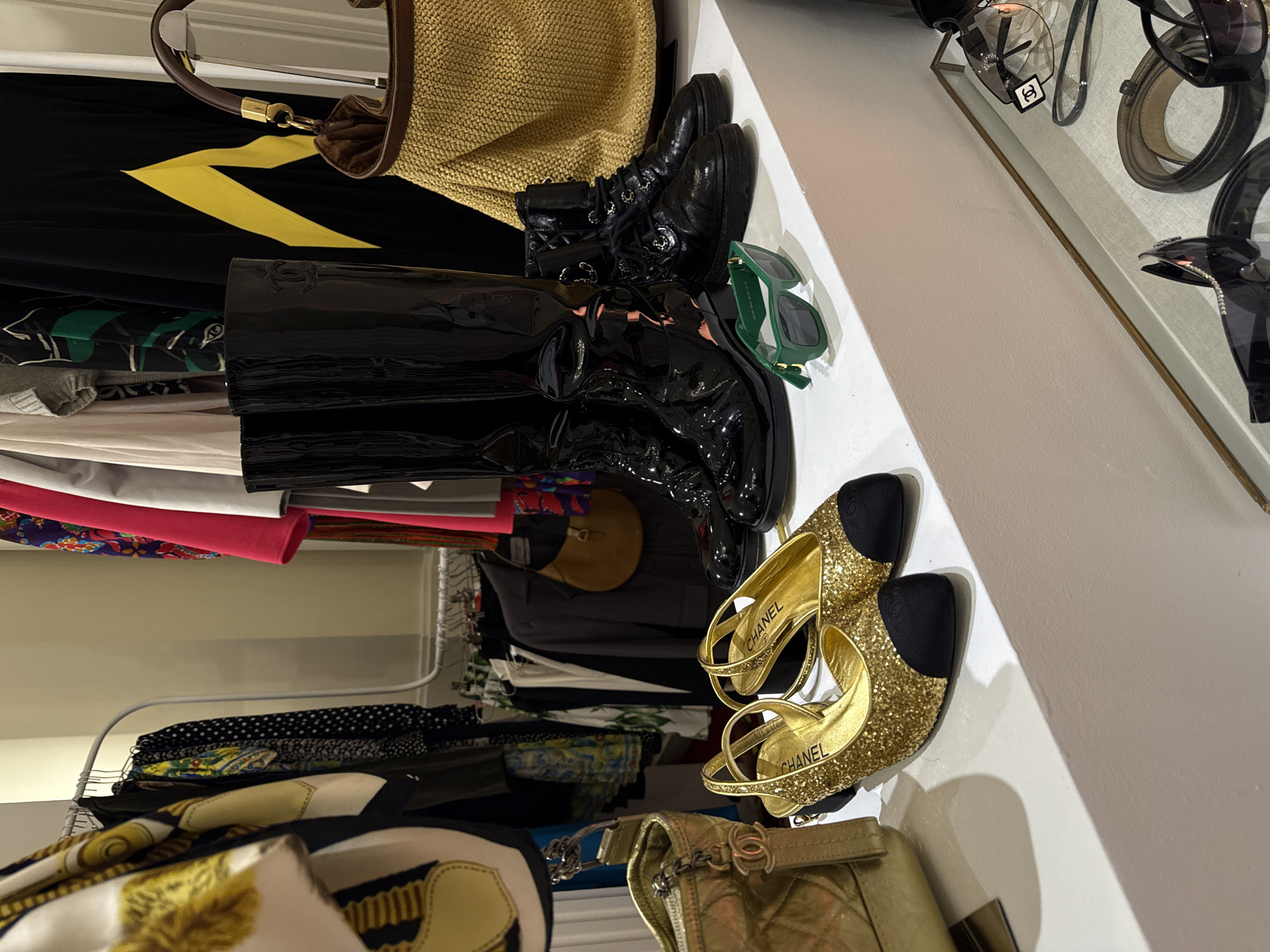 The image depicts a closet shelf containing fashion items. On the top shelf are two pairs of black boots (one combat style and one patent leather thigh-high), a pair of green sunglasses, a gold Chanel handbag, and two gold glitter and black cap-toe Chanel shoes. Clothes hang on racks in the closet. A straw tote bag with leather handles rests above the boots. The location is likely a private residence closet but is not definitively located in Mexico City. There are no people present in the scene.
