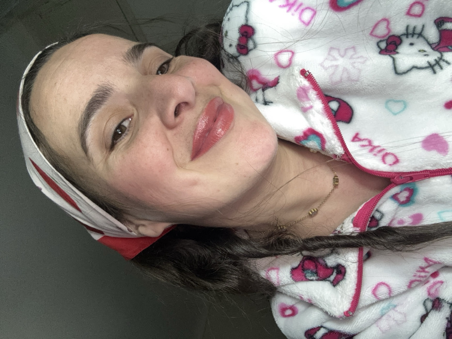 A woman with fair skin and brown hair tilted her head and smiled at the camera. She's wearing a red and white headscarf and a pink and white hooded jacket with a cartoon character pattern. A gold chain necklace is visible. Her makeup is natural, with glossy lips and rosy cheeks. The background appears to be a neutral wall, and the image is slightly rotated.