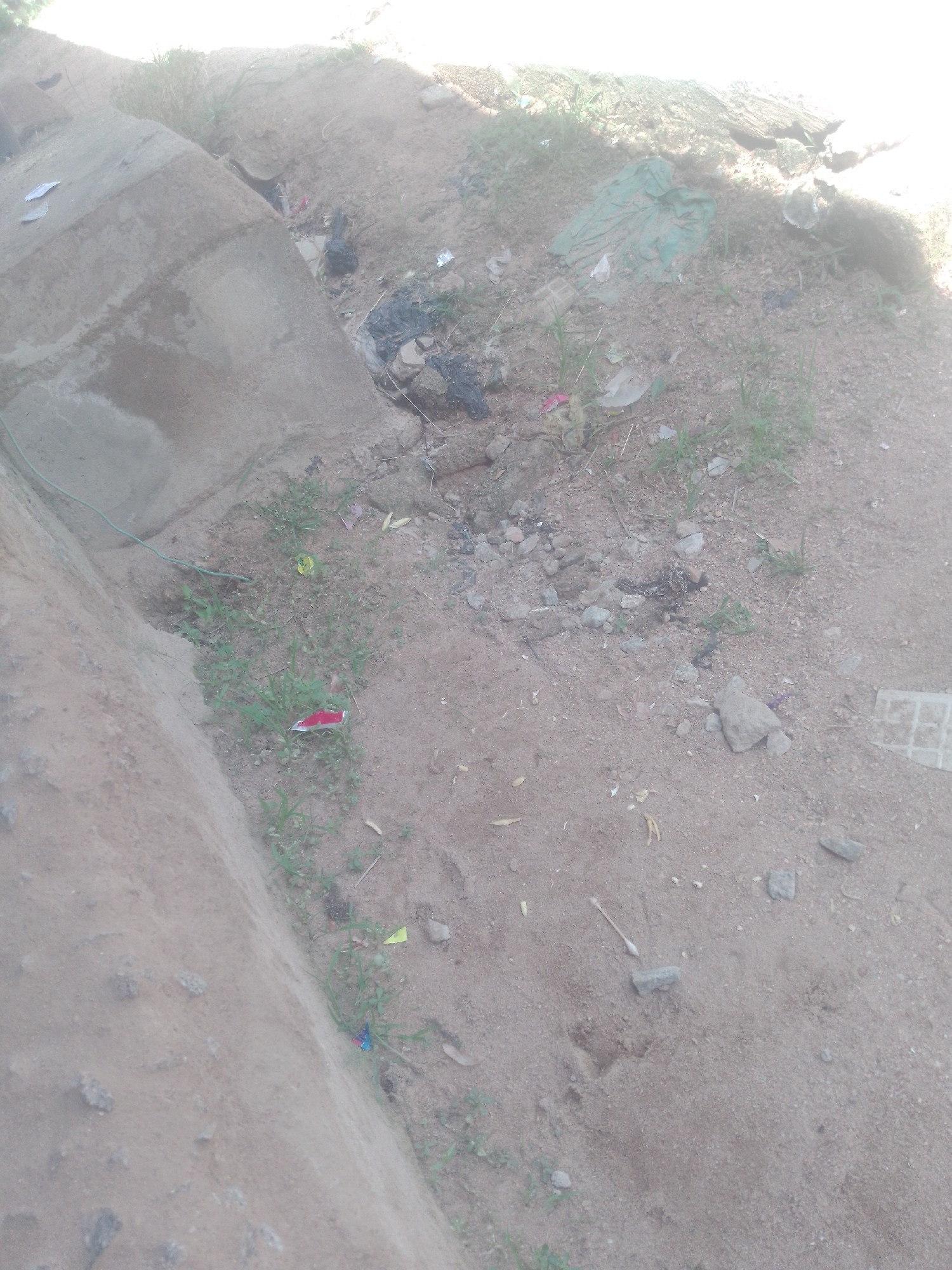 The image depicts an outdoor scene in Jauro Yinu, Nigeria, characterized by a dry, dusty ground littered with debris. A concrete structure, possibly a wall or curb, is visible on the left side of the frame, with a green wire running along its base. The ground is a mixture of sand and small rocks, with sparse patches of dry grass and weeds. Several pieces of trash are scattered across the scene, including what appears to be a crumpled green plastic sheet, black plastic bags, and smaller pieces of colorful wrappers. A small red and white object, possibly a candy wrapper or a piece of packaging, is noticeable among the vegetation. In the distance, the bright sunlight suggests an open area. There are no people or animals present in the image. The overall impression is one of an unkempt and neglected outdoor space.