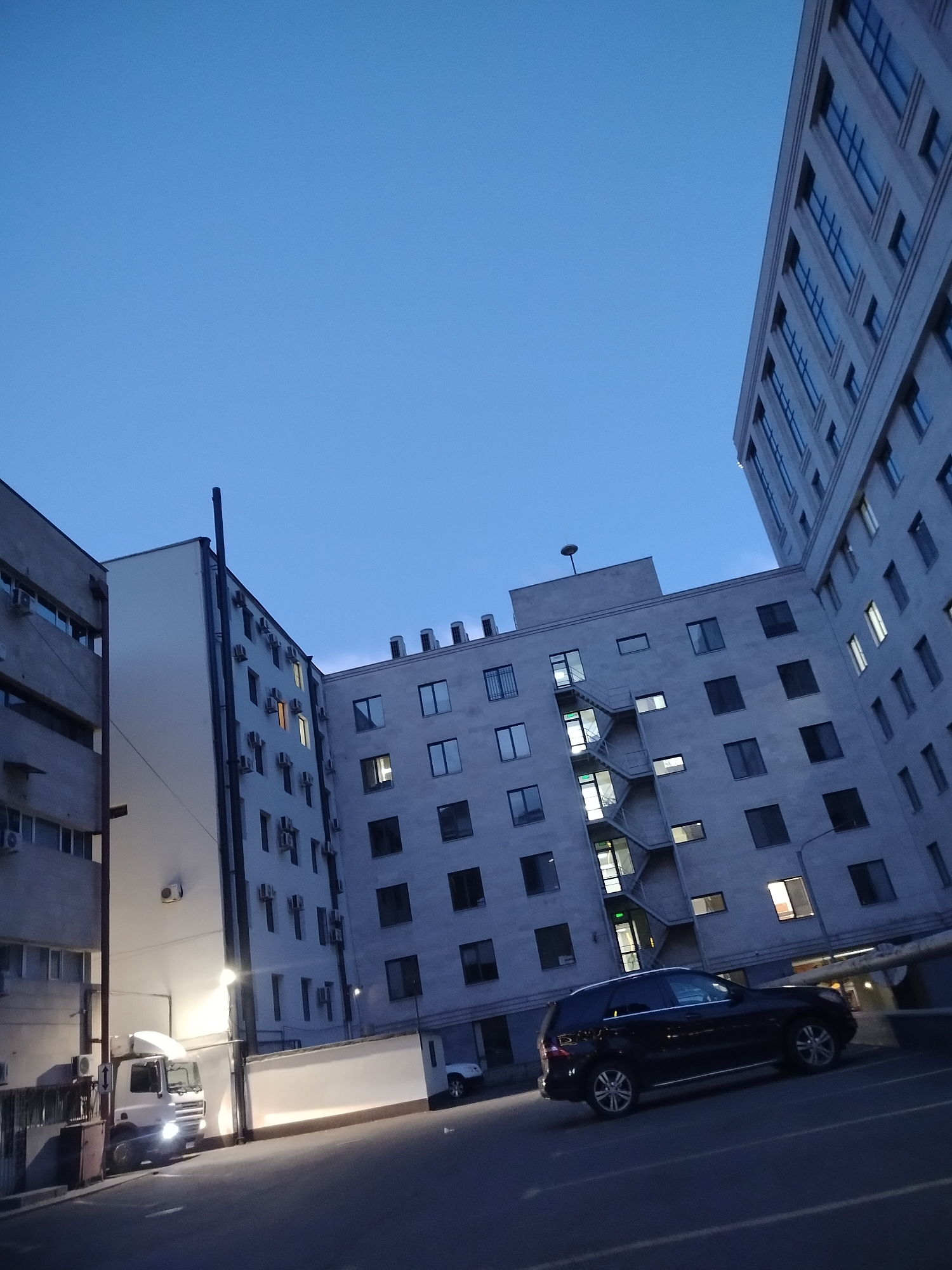 The image captures an urban scene in Yerevan, Armenia, at twilight or dusk. Several modern buildings with numerous windows dominate the frame. An external fire escape is visible on one of the buildings. A dark-colored SUV is parked on the street. A white truck is partially visible behind a low wall. The sky above is a deep blue. The street is dimly lit. No people are visible in the scene.