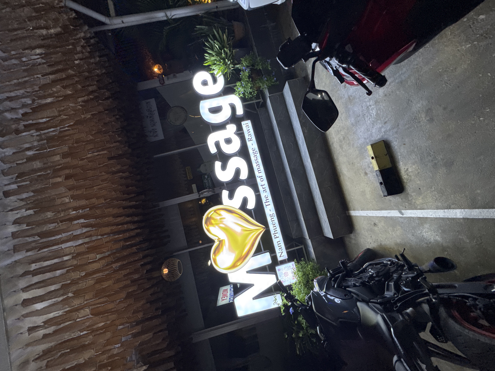 The image shows an angled view of the "Missage" massage parlor in Rawai, Thailand. The sign prominently displays the word "Missage" in large, lit-up white letters, with a golden heart-shaped symbol replacing the "i." Below, in smaller font, it reads "Nam Phueng - The art of massage - Rawai." A black and red motorcycle is parked in front of the entrance. The building's exterior has a brick-like textured wall. The location is near a parking area marked with white lines. A piece of vehicle maintenance equipment is also visible. A Thai language inscription, "น้ำผึ้ง นวดเพื่อสุขภาพ," is present on the establishment's facade. A round decorative light fixture hangs nearby.
