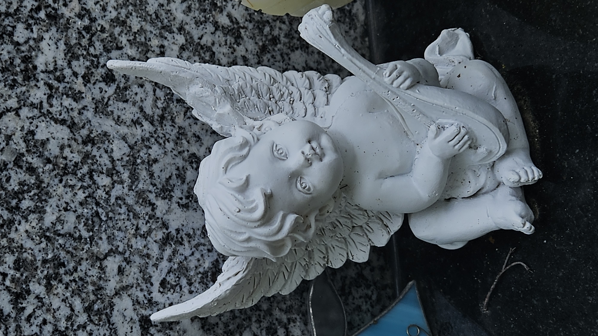 A white cherub statue sits on a dark polished surface. The cherub has wings and is holding a stringed instrument, possibly a lute, in its hands. Its head is tilted slightly to the right, and it appears to be looking forward. The background is a speckled grey granite surface. There is a small twig on the dark surface to the left of the cherub. The statue is likely a grave marker or memorial. Without further information or context, it is impossible to determine the specific location within Prešov, Slovakia.