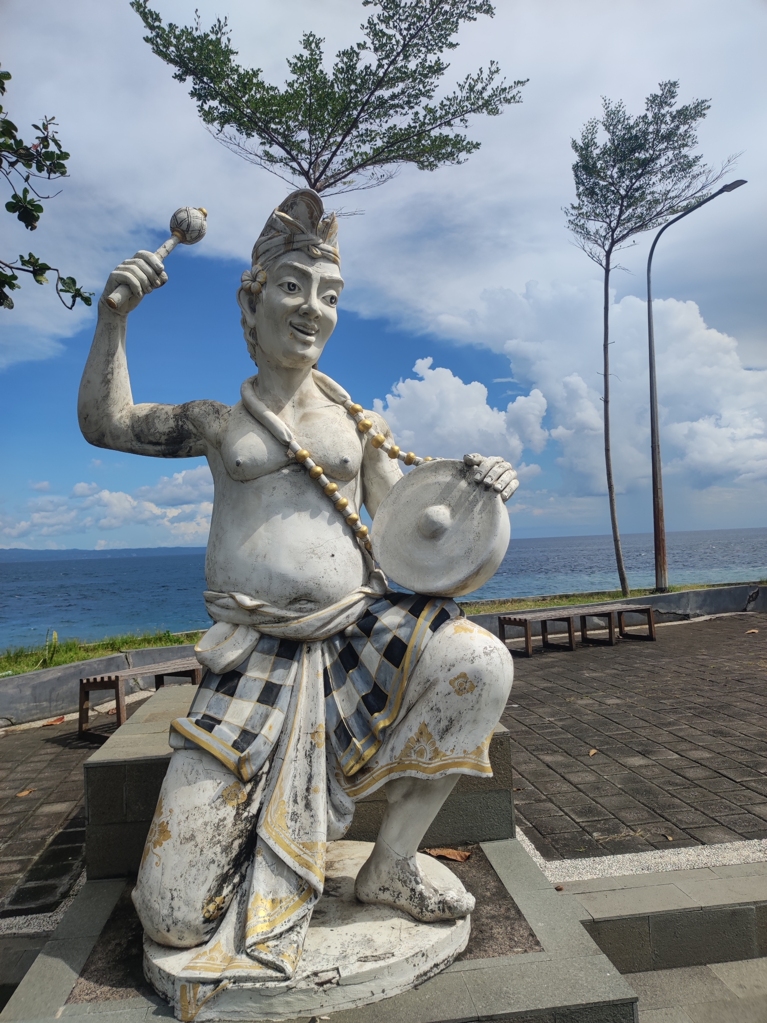 The image captures a weathered, white statue of a Balinese man in Padangbai, Indonesia. The statue is positioned on a paved terrace, with the ocean visible in the background. The man is depicted kneeling, holding a round, disc-shaped instrument in his left hand and a mallet-like object in his raised right hand. He is wearing a checkered fabric around his waist and a headdress. The sky is blue with scattered clouds. Trees flank the statue, one on either side. Benches are situated behind the statue. Two tall, thin poles, possibly light fixtures, are also visible near the edge of the terrace overlooking the ocean.