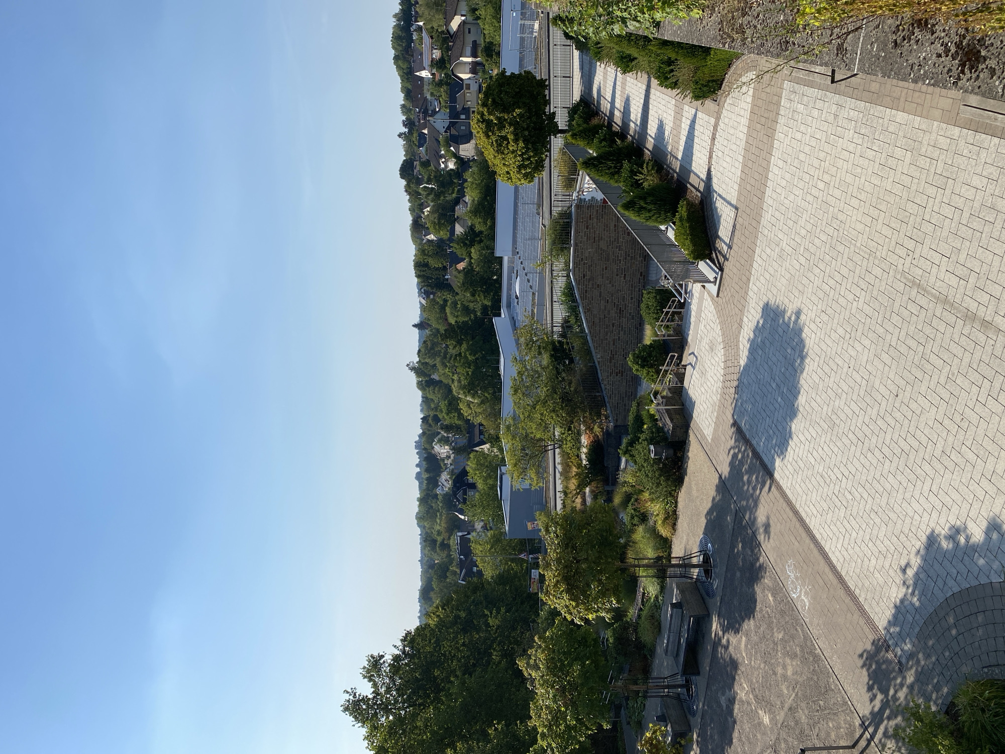 The image depicts a daytime view of a public outdoor area with a cityscape in the background, likely in Altenkirchen, Germany. The foreground is dominated by paved walkways and landscaped garden beds. In the left foreground, a paved area with some loose gravel is visible, featuring two benches and some small trees protected by circular grates. Adjacent to this is a section of walkway paved with rectangular blocks arranged in a herringbone pattern, bordered by a darker band of blocks.
Moving towards the center and right, the paving continues, with a distinct change in pattern to smaller rectangular blocks laid in a straight, interlocking pattern. This area is lined with a metal railing and various evergreen bushes and small trees. A stone wall with some ivy growing on it forms the right edge of the scene. A set of stairs with a metal handrail leads down from the upper level to a lower paved area, also bordered by railings and landscaping.
In the midground, there's a view of a modern building with a white roof structure and a blue-gray facade, behind which is a road with some signage. Beyond this, the background reveals a residential area with houses nestled amongst lush green trees, suggesting a suburban or semi-rural environment within the city. The sky is clear and bright blue, indicating sunny weather. There are no people visible in the image. The overall impression is of a well-maintained public space with a mix of natural and architectural elements.