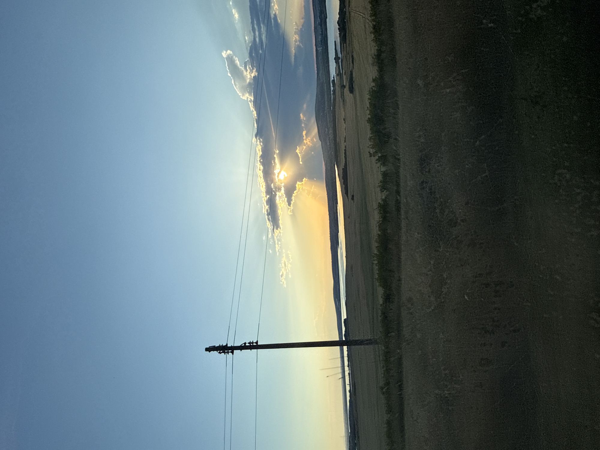 This image depicts a wide-angle view of a rural landscape at sunset, likely near Tvarditsa, Bulgaria. The sky is a gradient of blue, transitioning to warm yellow and orange hues near the horizon where the sun is setting behind clouds. The sun's rays are visible, piercing through the clouds and casting a dramatic effect on the scene. A single wooden utility pole with wires stands prominently in the left mid-ground, aligned with power lines that stretch across the frame. In the background, there is a body of water, possibly a lake or reservoir, bordered by rolling hills and vegetation. The foreground is dominated by fields of dry grass and foliage, suggesting a late summer or early autumn setting. The overall impression is one of natural beauty and tranquility, characteristic of the Bulgarian countryside.