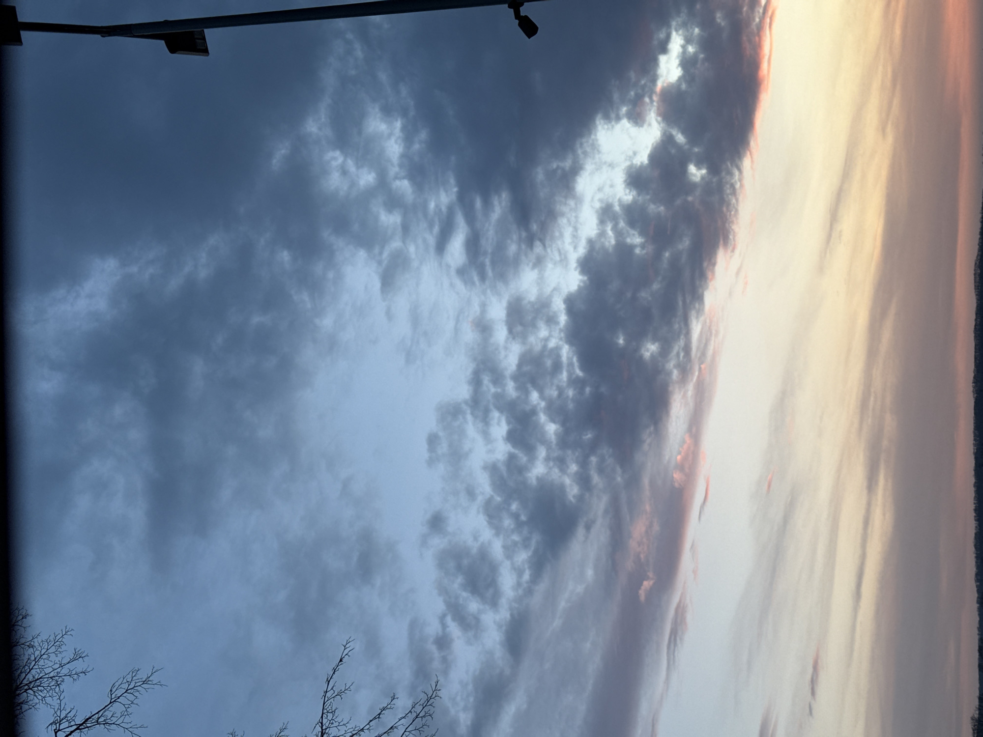 The image shows a sky with a mix of dark and light clouds. Some branches of trees are visible in the lower left corner. A light post with a fixture is visible in the top left corner. The clouds on the right side of the image are illuminated with a yellow/orange color. The photograph may have been taken in Votkinsk, Russia, though there is no clear indication this is the location from the image itself.